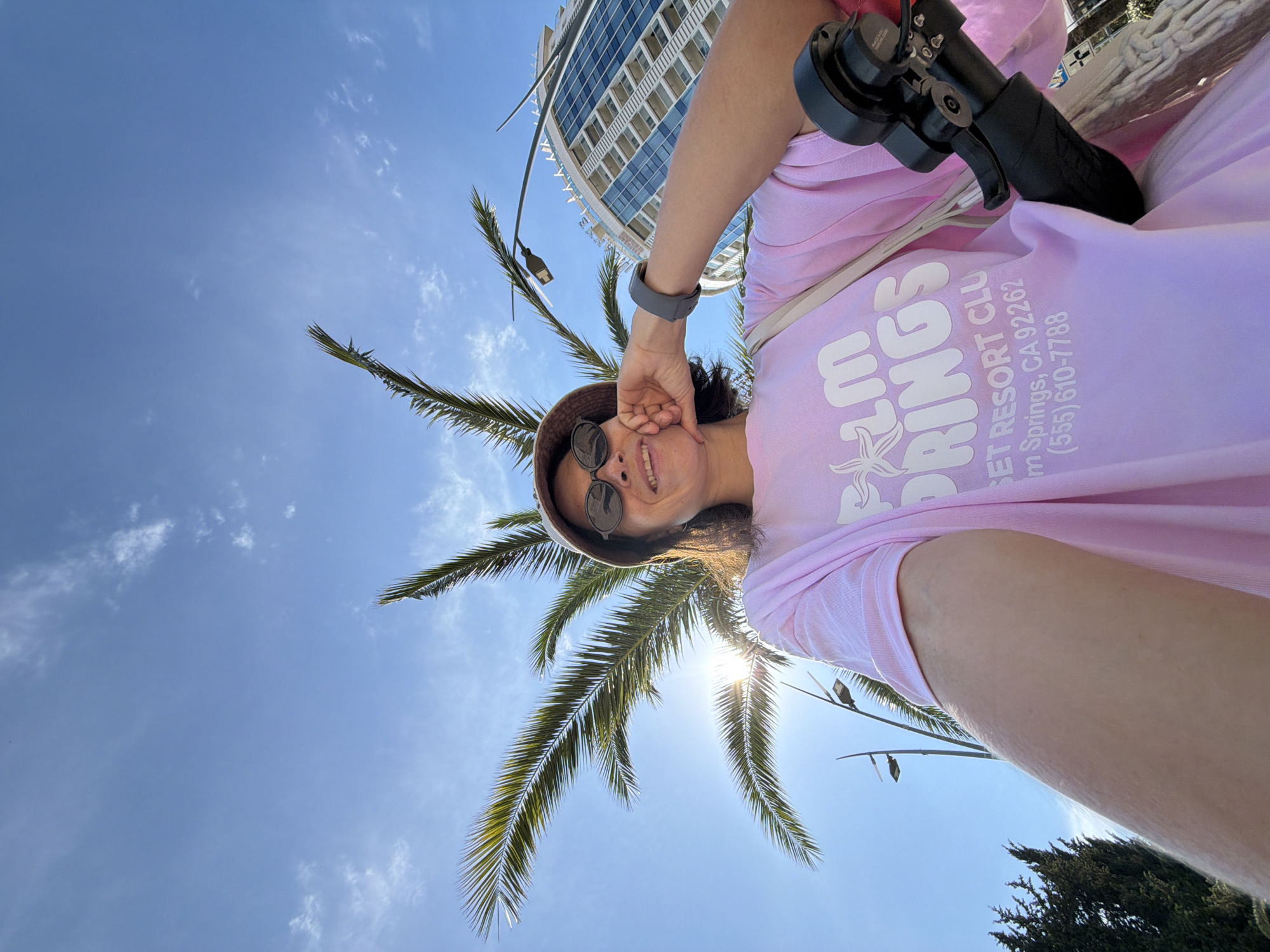 The image shows a woman in a pink t-shirt and a bucket hat taking a selfie outdoors in Budva, Montenegro. She is wearing sunglasses and resting her hand on her chin. Behind her, a palm tree is visible against a clear blue sky with some wispy clouds. A modern building with glass windows and balconies is also in the background, along with streetlights. The woman is holding a device that appears to be a scooter or bicycle handle. The text on her t-shirt reads "Palm Springs Resort Club, Palm Springs, CA 92262, (555) 610-7788".