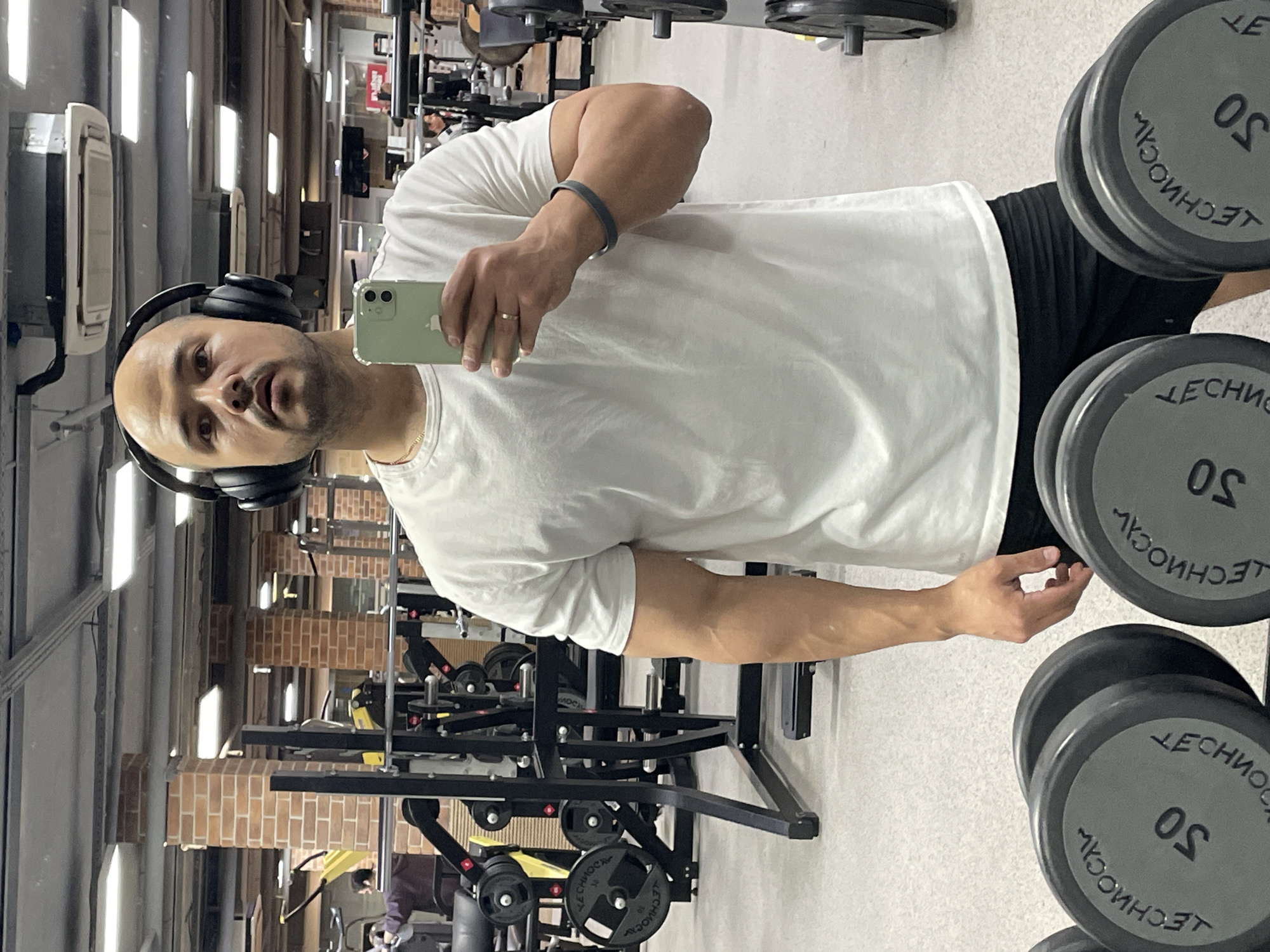 A bald man wearing headphones and a white t-shirt is taking a selfie in what appears to be a gym. He is holding a light green phone in his right hand. The background includes weightlifting equipment and dumbbells. Weights are labeled "TECHNOGYM" and are 50 kg. The lighting suggests an indoor environment with multiple light sources. A partial view of a brick wall can be seen in the background, further implying an interior setting. It is not possible to determine if the location is in Izhevsk, Russia, based on this image.