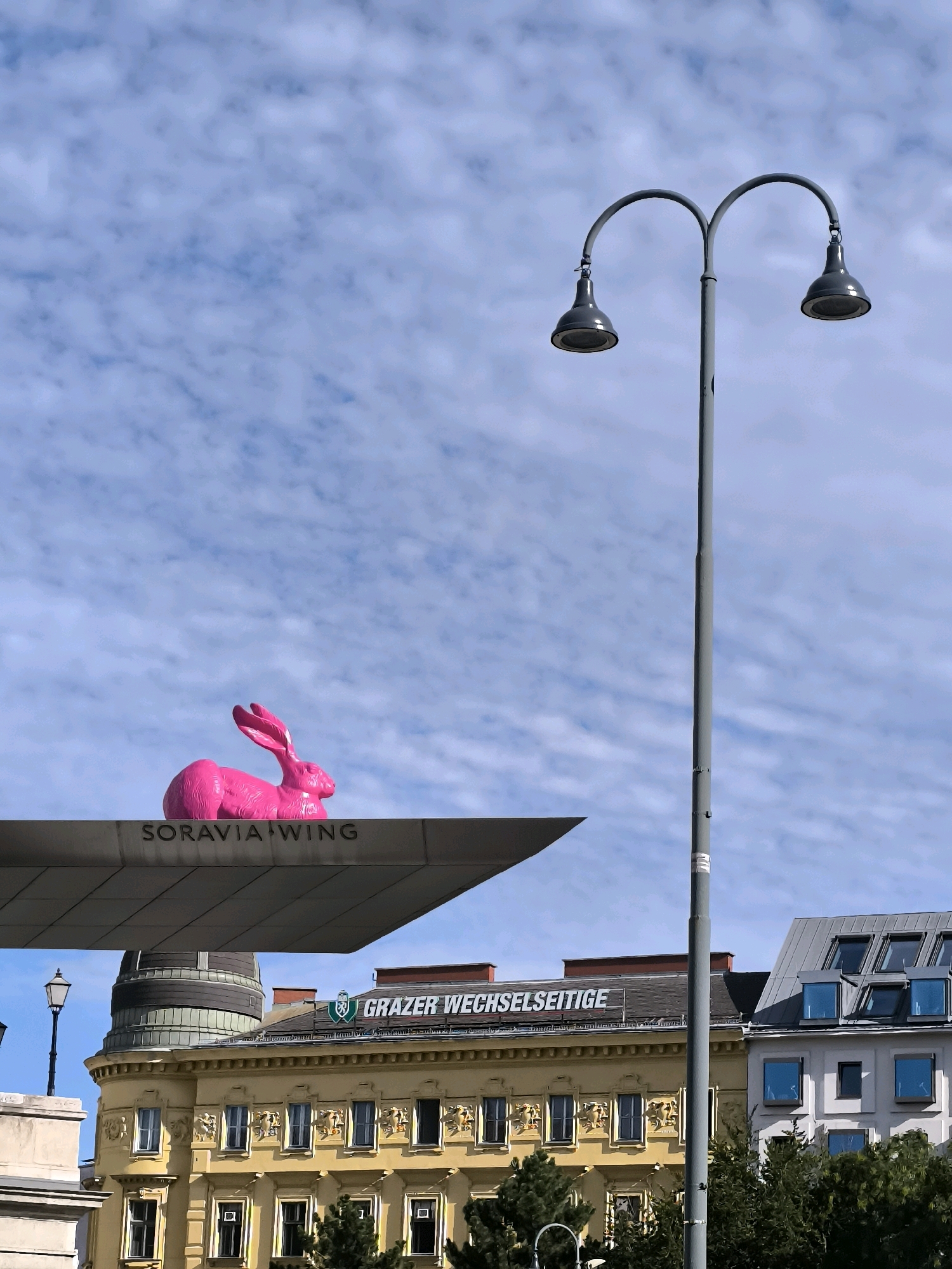 A large, bright pink rabbit sculpture is perched on the edge of a modern, grey structure labeled "SORAVIA WING." The structure juts out over a historic yellow building with ornate architectural details and numerous windows. Above the yellow building, the word "GRAZER WECHSELTSEITIGE" is visible on a dark sign. To the right of this building is a lighter-colored building with a grey, sloped roof and dormer windows. A tall, grey street lamp with two lights stands prominently in the foreground, positioned to the right of the rabbit sculpture. The sky above is blue with scattered, puffy white clouds. The overall scene appears to be an urban setting in Vienna, Austria, likely a public space that combines modern art installations with historical architecture. There are no people visible in the image.