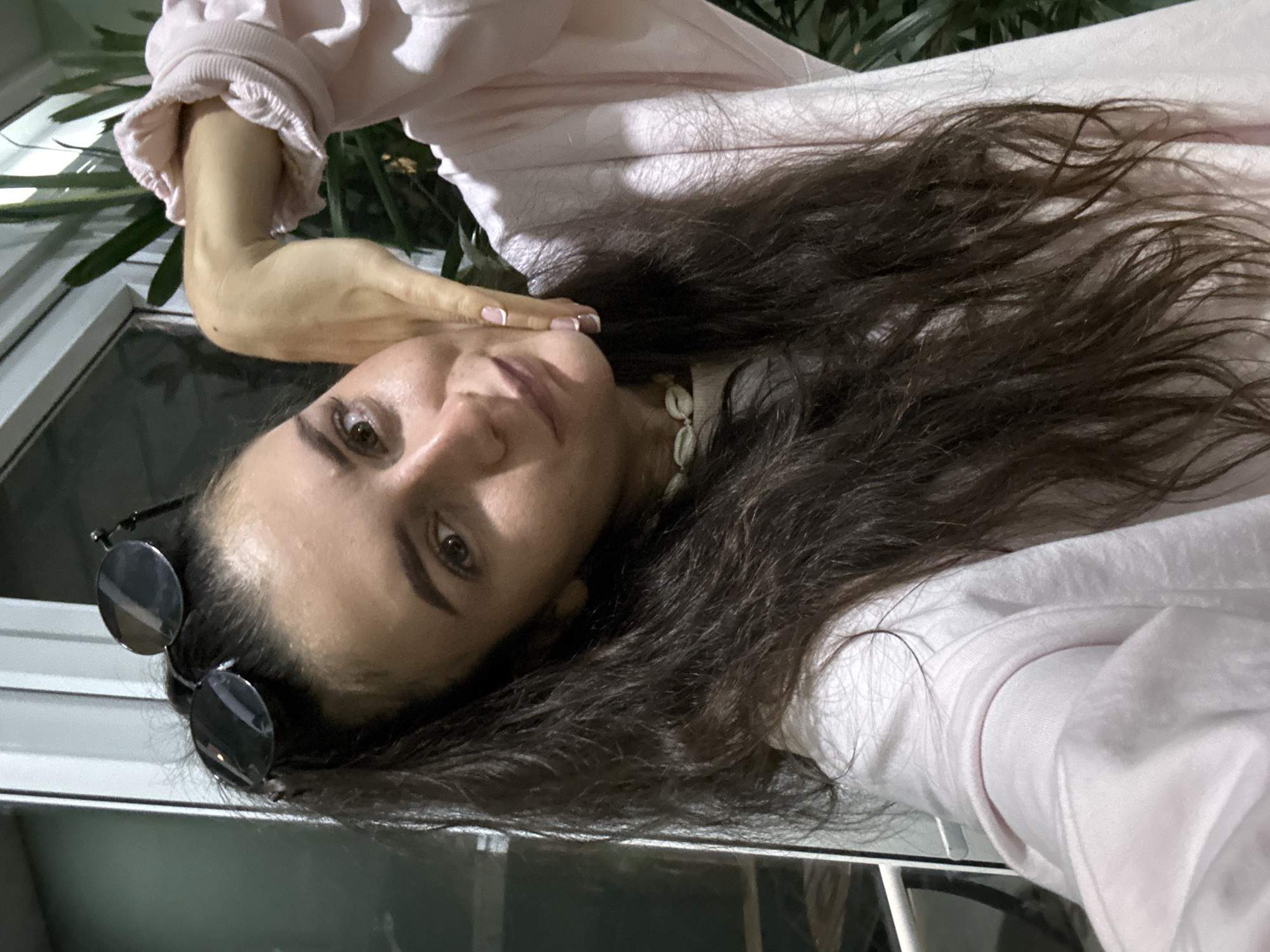 A woman is pictured in a close-up, selfie-style image. Her long, dark hair cascades around her face. She wears a light pink sweater and a shell necklace. Sunglasses are perched on her head. Her right hand is gently touching her face near her chin. The location is identifiable as Da Lat, Vietnam, due to the architectural style and foliage visible in the background. The setting appears to be an outdoor area, possibly a patio or balcony.