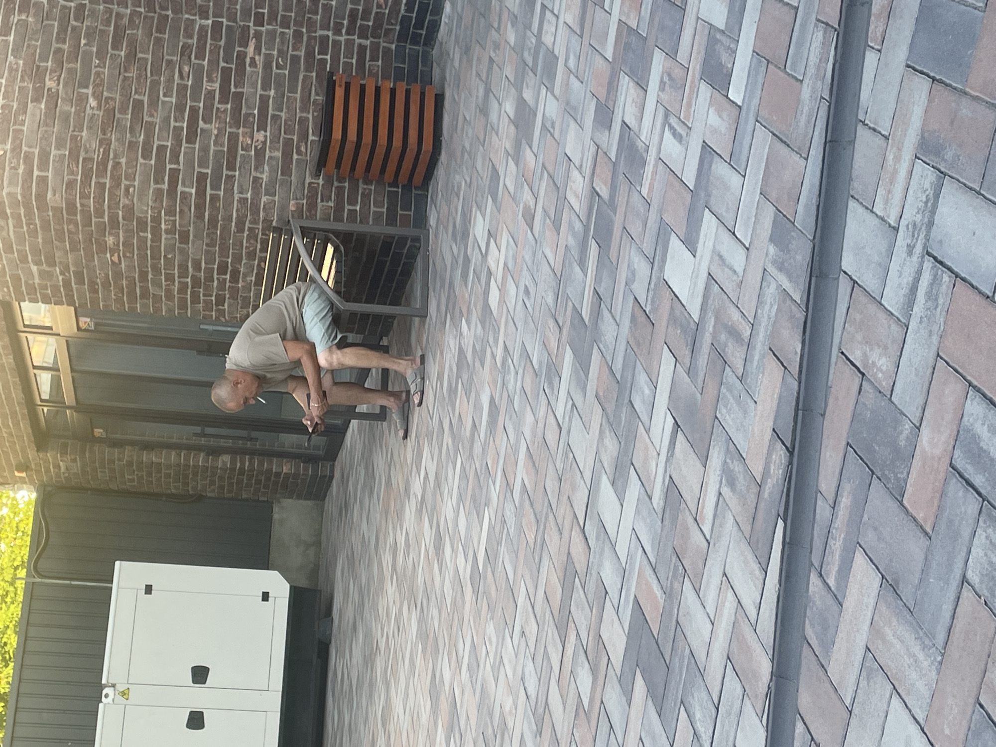 A bald, middle-aged man in a light brown t-shirt and light blue shorts is sitting on a dark metal bench with a slatted wood seat. He is holding a mobile phone in his left hand and a cigarette in his right hand, with the cigarette burning and producing smoke. He is looking down at his phone. The bench is situated on a paved patio made of interlocking rectangular stones in various shades of grey, brown, and pink. Behind the bench is a brick wall with dark mortar. To the left of the bench is a large white generator with a black base and two black doors. Further to the left is a dark grey metal fence and lush green trees. To the right of the bench is a trash receptacle with horizontal orange and black slats. The scene appears to be outdoors in a courtyard or patio area.