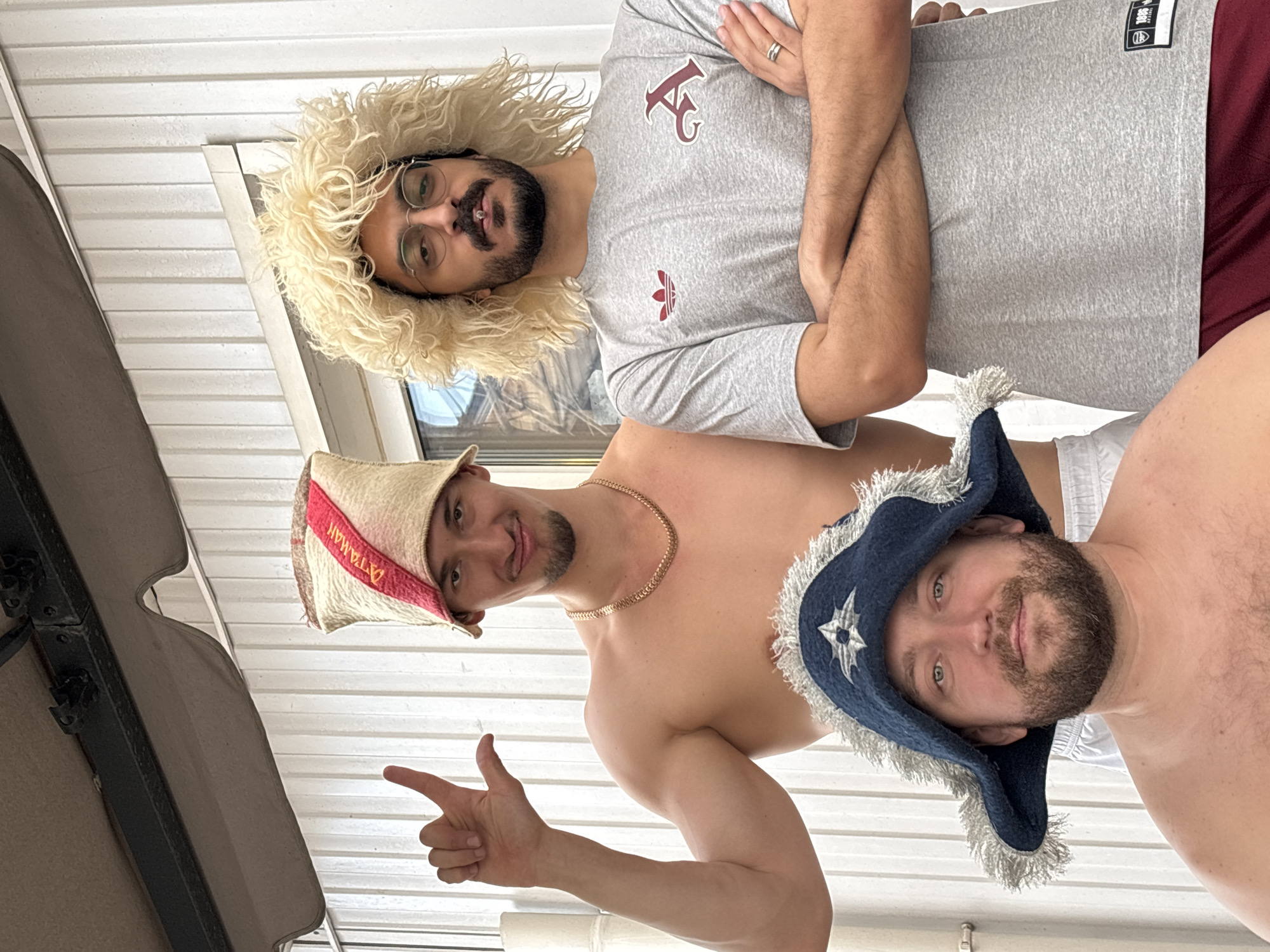 Three men are posing for a photo. The man at the top is wearing a gray t-shirt with a logo and a large, curly white hat. He has a beard and glasses. The man in the middle is shirtless and wearing a hat with "ATAMAN" written on it. He is also wearing a gold chain and pointing. The man at the bottom is shirtless and wearing a blue hat. The background is a white, corrugated surface. It is not possible to identify the location as Krasnoyarsk, Russia from the provided image.