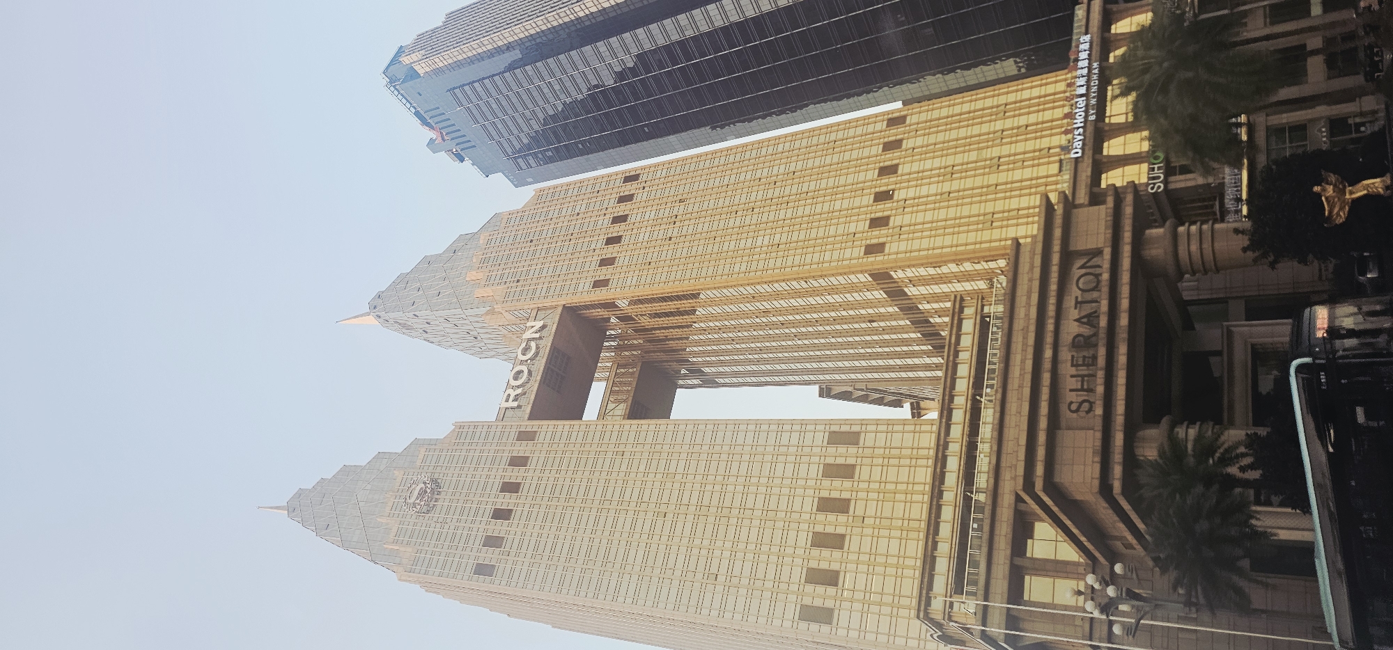 The image shows a low-angle view of the Sheraton hotel in Nan'an District, China. The hotel is a prominent golden skyscraper with a distinctive stepped, pyramid-like roof structure. A connecting bridge spans between two sections of the building, with the letters "ROCN" visible on it. To the right of the golden skyscraper, a dark, modern glass skyscraper rises higher, reflecting the sky. In the foreground, the entrance to the Sheraton hotel is visible, featuring large pillars and the hotel's name in prominent lettering. A bus is positioned in front of the entrance, and palm trees and landscaping are present around the base of the buildings. The sky above is a pale, hazy blue, suggesting it might be early morning or late afternoon.