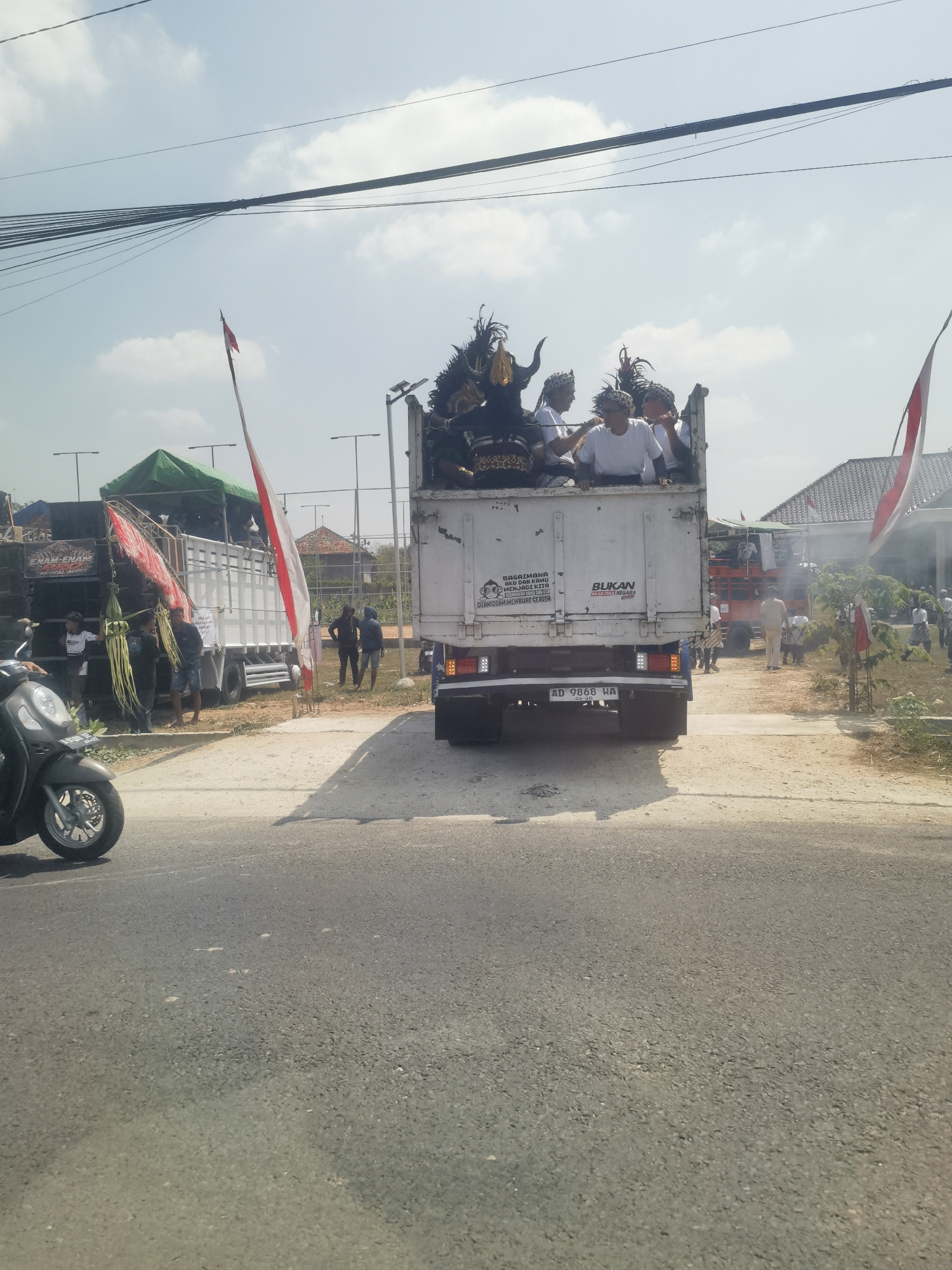 The image captures a scene on a street in Tambaksari, Indonesia, with a convoy of trucks and people. The foreground shows a paved road with a motorcycle on the left. A white truck is the central focus, carrying several individuals dressed in white tops and traditional headwear. The truck also has large decorative figures, possibly part of a parade or cultural event, with dark colors and prominent horns and feathers. The truck's rear displays a license plate reading "AD 9868 HA".
Behind the white truck, another truck is visible, also with people inside. To the left of the white truck, a separate truck is heavily laden with sound equipment and speakers, adorned with red and white banners and green foliage. Several people are gathered around this sound truck.
The environment includes power lines overhead and a bright sky with scattered clouds. In the background, there are buildings with tiled roofs, a green tent, and several lamp posts. Indonesian flags are prominently displayed, including one on a tall pole near the white truck and smaller ones on buildings in the distance. The overall impression is that of a public gathering or procession taking place outdoors.