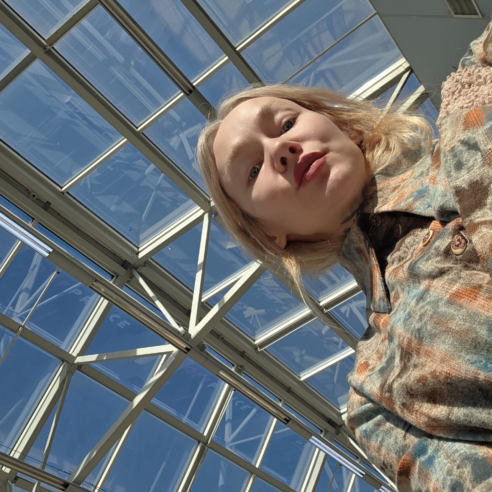 The image shows a woman with fair skin, blond hair, and blue eyes looking down at the camera. She is wearing a patterned button-up shirt with a high collar. The background is a glass roof structure with a grid of white supports, and a clear blue sky is visible through the glass panels. The location is likely a shopping mall or atrium-like space in the city of Saint Petersburg, Russia, as indicated by the partial text "PA CE" visible on one of the glass panels.