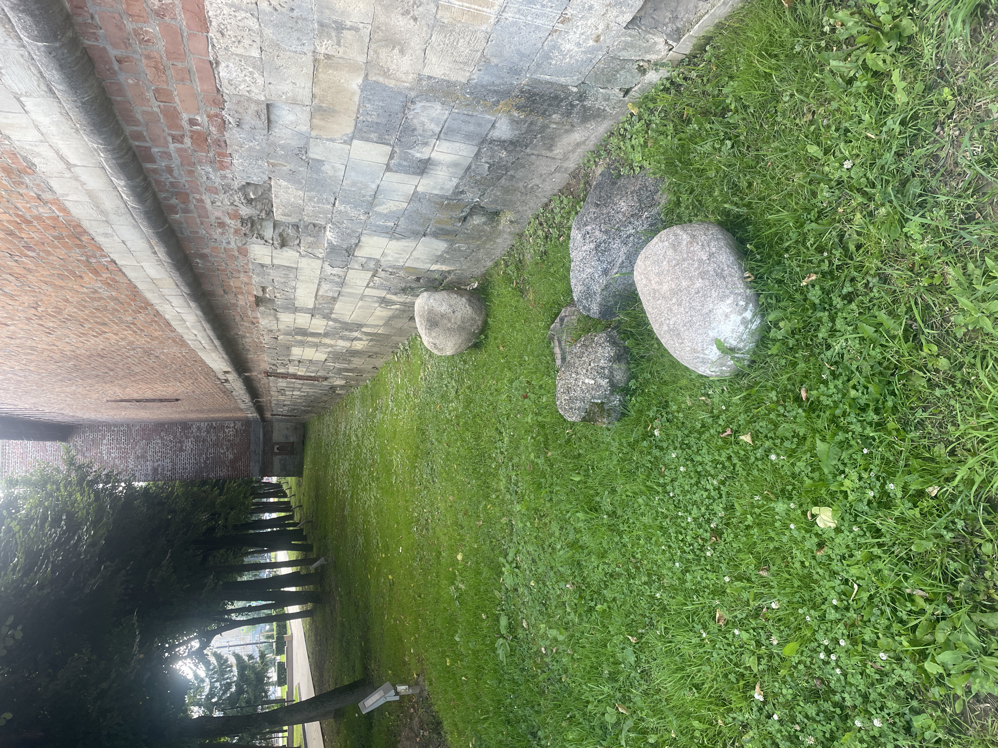 The image shows a section of a brick wall with a stone foundation in Sof'ino, Russia. The wall is weathered and shows signs of age. In front of the wall is a grassy area with several large rocks scattered on the ground. A line of trees with dense green foliage can be seen to the left of the wall, casting shadows on the grass. The overall environment appears to be an outdoor public space, possibly a park or the grounds of a historic building.