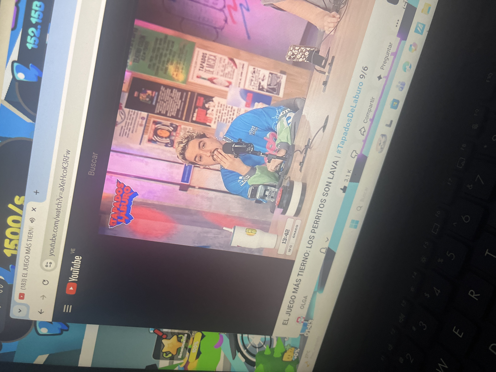 The image shows a laptop screen displaying a YouTube video. The video features a man with blonde, styled hair, wearing a blue long-sleeved shirt, speaking into a microphone. He is seated at a table with other microphones and a white cup. Behind him, the wall is decorated with posters. The YouTube interface is visible, with the video title "EL JUEGO MÁS TIERNO: LOS PERRITOS SON LAVA | #TapadosDeLaburo 9/6" displayed. The URL bar shows "youtube.com/watch?v=aXeHcoK3RFw". To the left of the screen, a game interface with graphics and text like "Renacimiento" and percentages is partially visible. The keyboard of the laptop is also visible in the foreground. The scene is indoors, likely a room where the laptop is being used to view the YouTube video. There is no information in the image that can be used to determine a location in Santa Ana, Venezuela.