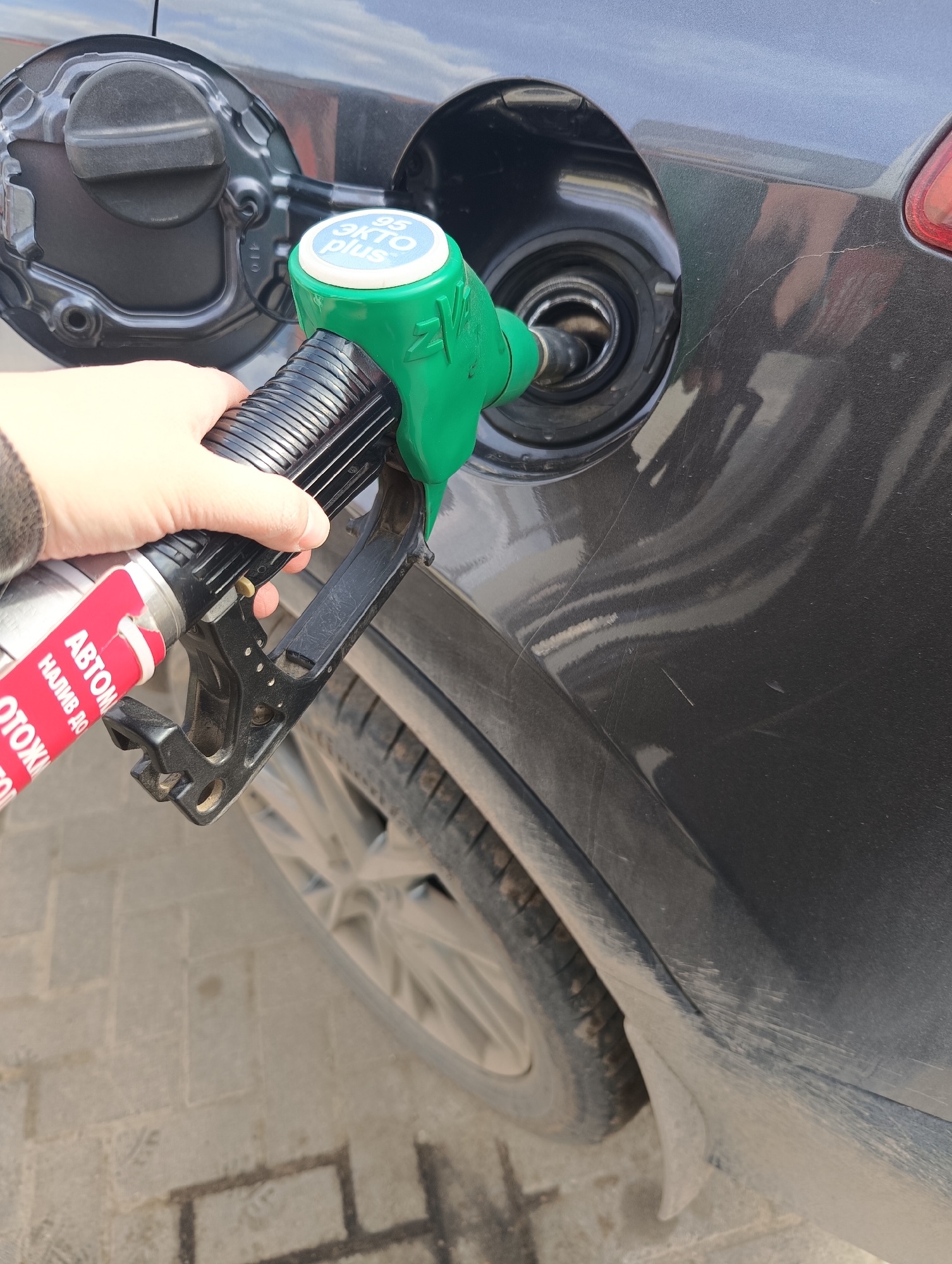 A person is seen refueling a dark-colored car at what appears to be a gas station during the daytime. A light-skinned hand, possibly female, holds a green and black fuel pump nozzle. The nozzle is inserted into the car's open fuel tank opening, which is located behind a black fuel cap that is swung open.
The fuel nozzle has a light blue and white cap with the text "95 ЭКТО plus" (95 EKTO plus) visible. A red and white label on the nozzle's handle displays Cyrillic text: "АВТОМАТ. НАЛИВ ДО ОТОЖИ...", which translates roughly to "AUTOMATIC. FILL UNTIL AUTO-SHUTOFF..." or "AUTOMATIC. FILL UNTIL PRESS...".
The car itself is dirty, with dust and grime evident on the lower body panels, wheel well, and tire. Numerous scratches and scuff marks are visible on the car's paint, particularly near the fuel cap area and on the rear fender. A faint "110" is partially visible inside the fuel cap area. The ground around the car is paved with light-colored stone or concrete blocks. The weather appears clear with bright, even lighting.