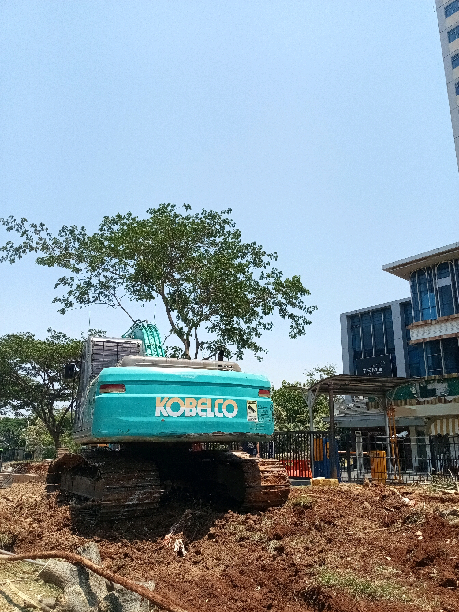 In Bekasi, Indonesia, a large, teal Kobelco excavator is situated on a construction site. The excavator's tracks are embedded in disturbed brown soil, suggesting recent digging or earthmoving activities. Several large tree branches, some with leaves, are visible both above and behind the excavator. In the background, a modern building with blue-tinted windows and white accents stands under a clear blue sky. A smaller structure with a covered awning and a black sign reading "THE TEMU COFFEE & EATERY" is also present. The overall scene depicts an active construction area within an urban environment.