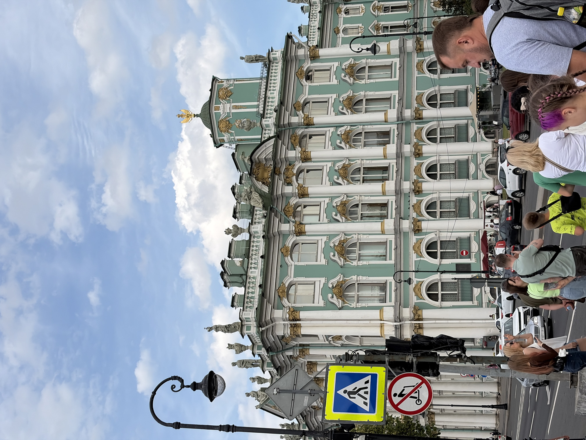 This is a daytime, eye-level, full-shot photograph of the Winter Palace in Saint Petersburg, Russia. The palace is a large, ornate building with light green walls, white columns, and gold accents. It is topped with statues and a golden eagle. In the foreground, there is a street with cars and several people waiting at a crosswalk. There are traffic lights and pedestrian crossing signs visible. The sky is blue with white clouds.