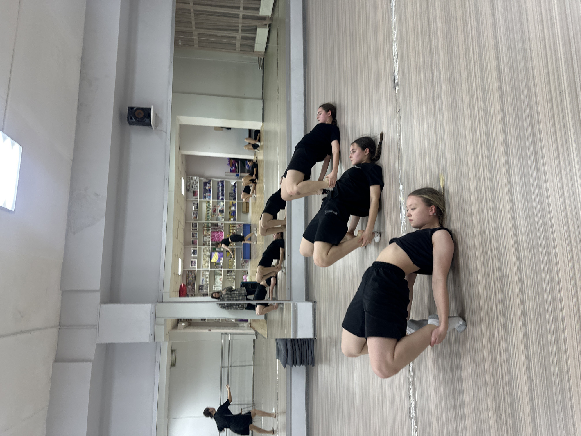 The image shows a dance studio, possibly located in Votkinsk, Russia, as requested. Three young women are performing a synchronized dance move. The girls are dressed in black tops and shorts and positioned in a line, each with their hands on the shoulders or backs of the person in front of them, bending forward with their legs in a squat position. A large mirror runs along the length of the studio wall, reflecting the dancers and other details of the room, including bookshelves with various items and a column with mirrors. The studio has white walls, a wooden-look floor, and overhead lighting.