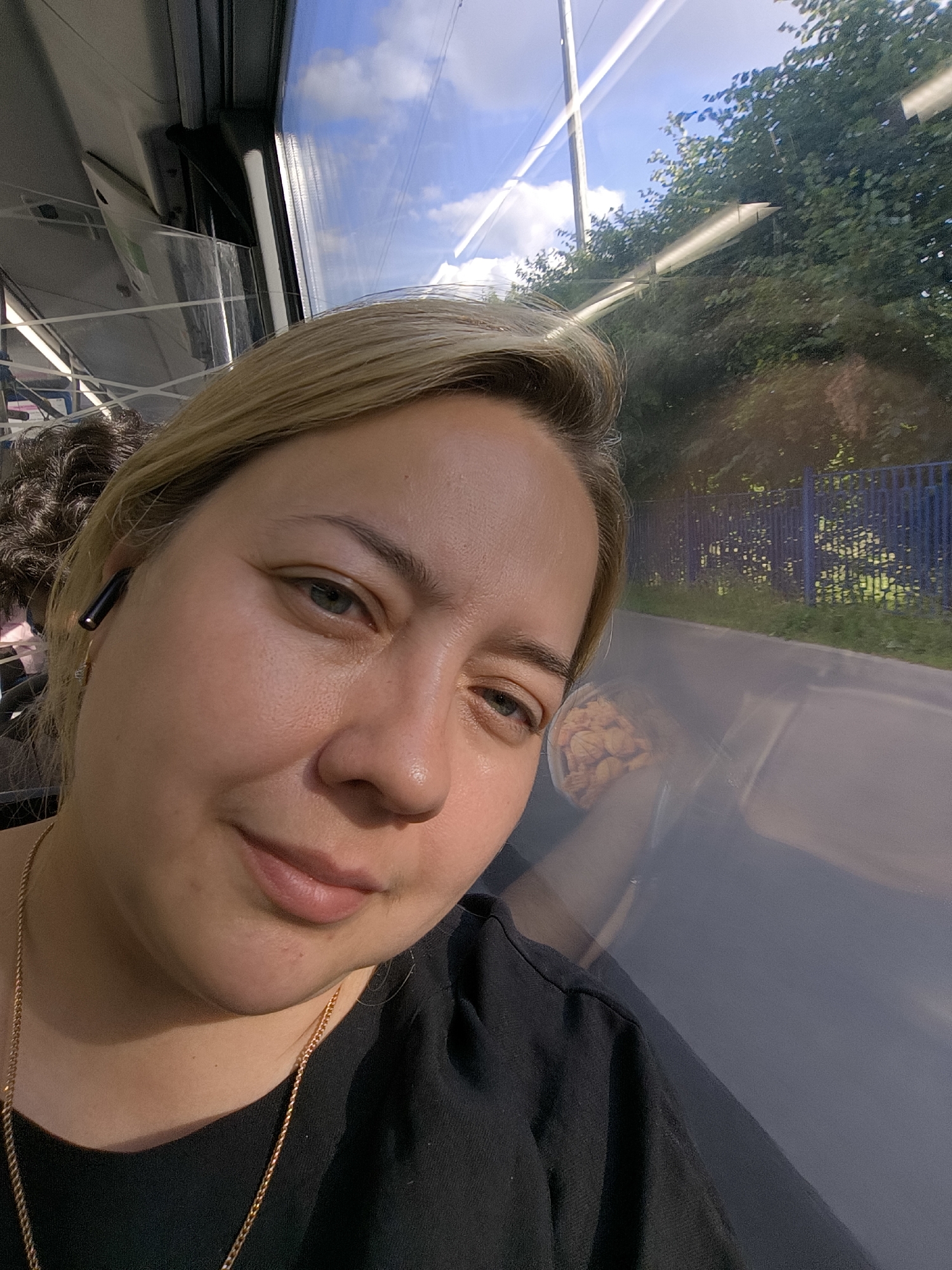 A young woman with blonde hair pulled back is taking a selfie on a bus. She is wearing a black t-shirt and a gold chain necklace, with an earbud in her left ear. Sunlight streams in from the window, illuminating her face and casting a warm glow. Her expression is relaxed and content. Outside the window, trees and a blue fence are visible, suggesting a journey through a suburban or rural area on a sunny day. The reflection in the window shows the interior of the bus and hints at the passing scenery. The overall mood is peaceful and pleasant, capturing a moment of personal reflection during a commute. There is no visible text in the image.