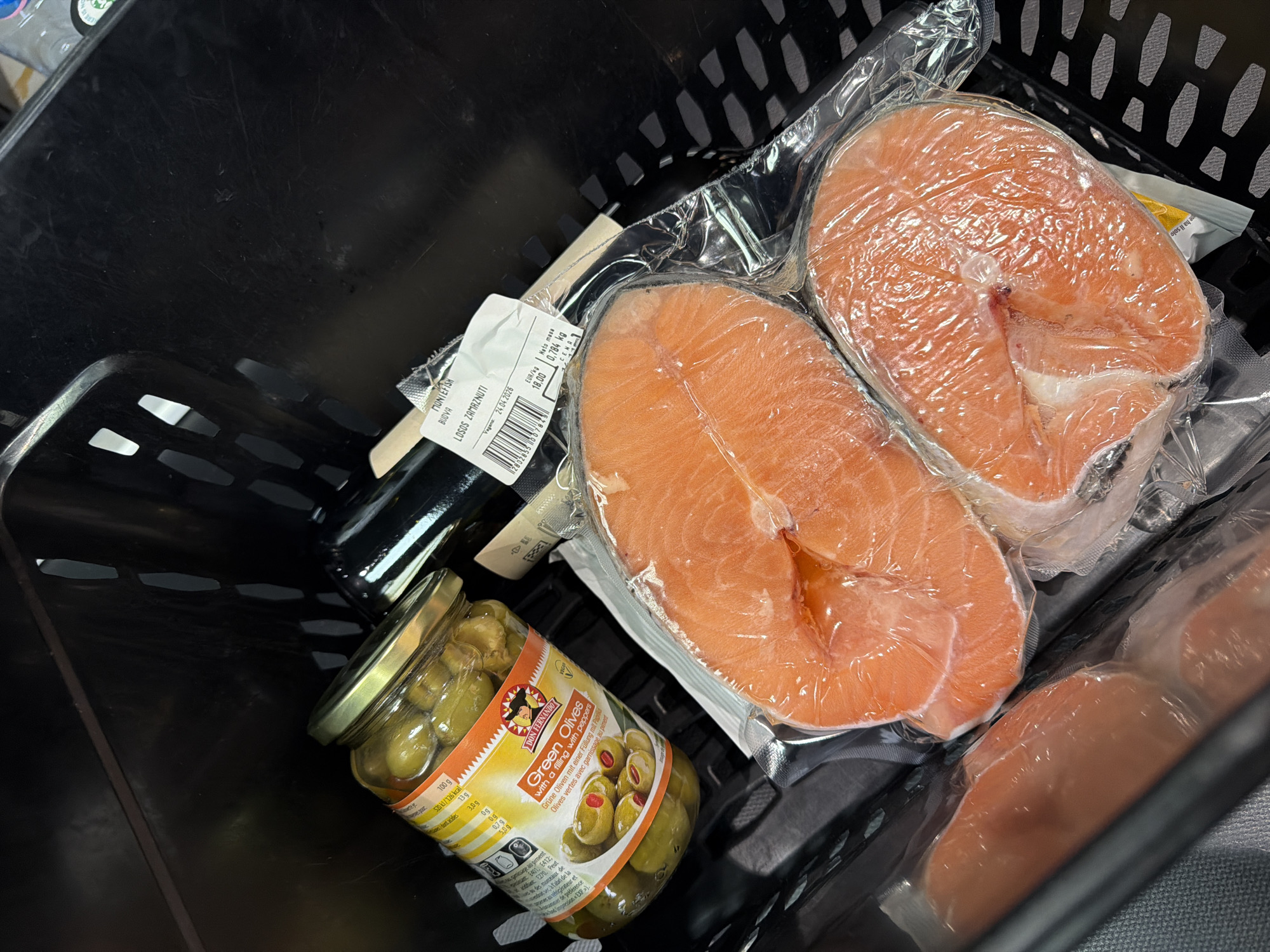 This image presents a close-up, overhead view of groceries situated within a black plastic shopping basket. The main subjects are two large, vibrantly orange-pink salmon steaks, individually vacuum-sealed in clear plastic. Beside the salmon is a glass jar of "DON FERNANDO Green Olives with a filling with peppers." A dark-colored bottle, possibly wine or a similar beverage, is partially visible on the left side of the basket.
A white product label is attached to the salmon packaging. Visible text includes "MONTEFISH," "BUDVA," and "LOSOS ZAMRZNUTI," which translates to frozen salmon. The label also specifies a net mass of "0.784 kg," a price of "EUR/kg 18.00," and a vegan date (likely expiration) of "24.04.2026," along with a barcode. The olive jar's label displays nutritional information, such as "520 kJ/126 kcal per 100 g," and descriptions in English, German, and French.
The scene depicts items typically purchased during a grocery trip. The "BUDVA" marking on the salmon label connects the context to Budva, Montenegro, suggesting the location of purchase. The items are packed within a textured black basket, under indoor lighting. No individuals are visible in the image.