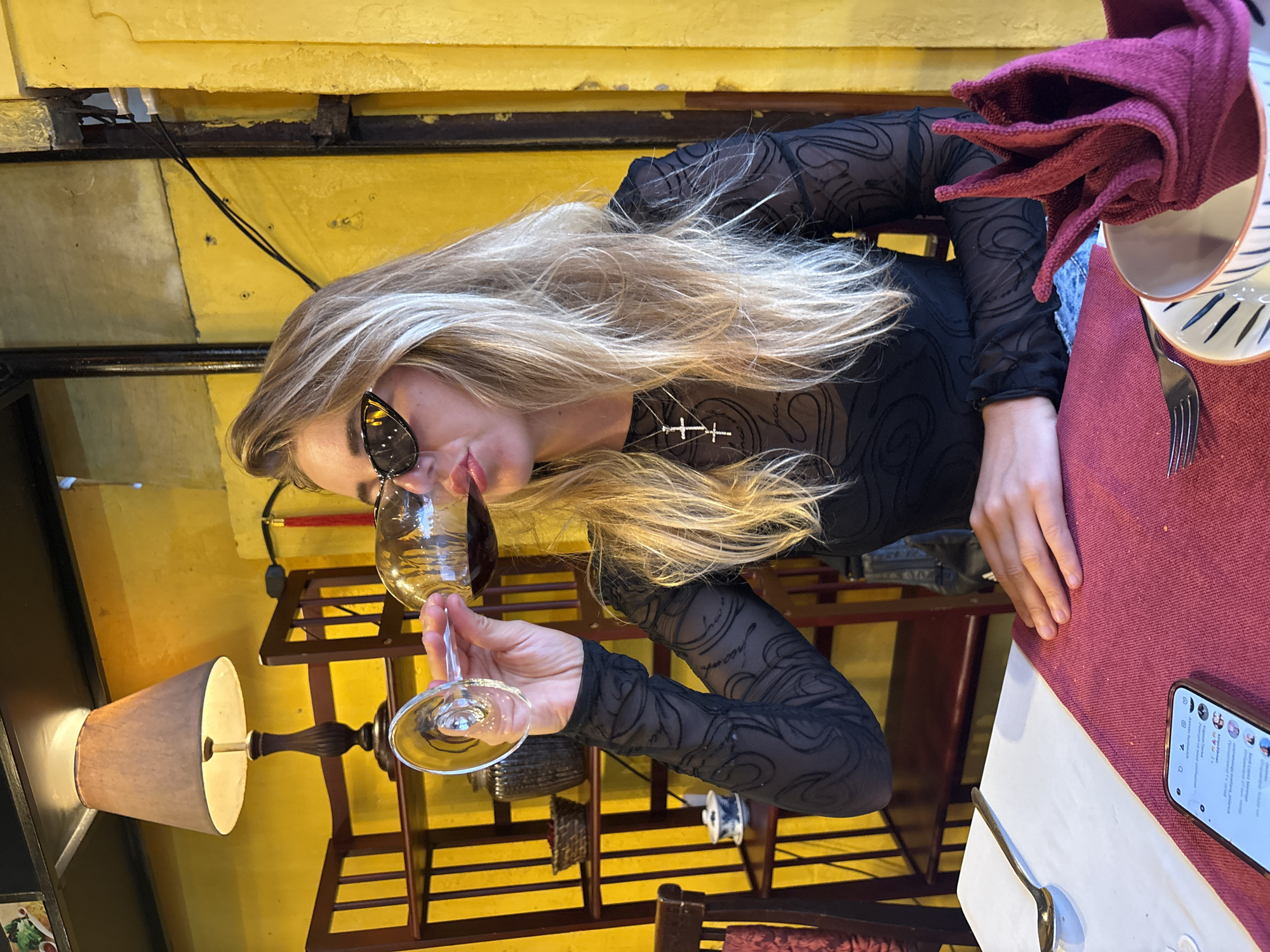 A fair-skinned blonde woman with dark sunglasses, wearing a sheer black top, sits at a table in what appears to be a restaurant or café. She is holding a glass of red wine to her lips, seemingly taking a sip. The table is set with a red tablecloth, white plates, silverware, a folded dark red napkin, and a cell phone. Behind the woman, there is a yellow wall with various pieces of furniture or shelving. In the lower left corner, there is a lamp with a light-colored shade. The overall setting suggests an intimate or casual dining environment. The location is Hội An Ward, Vietnam.