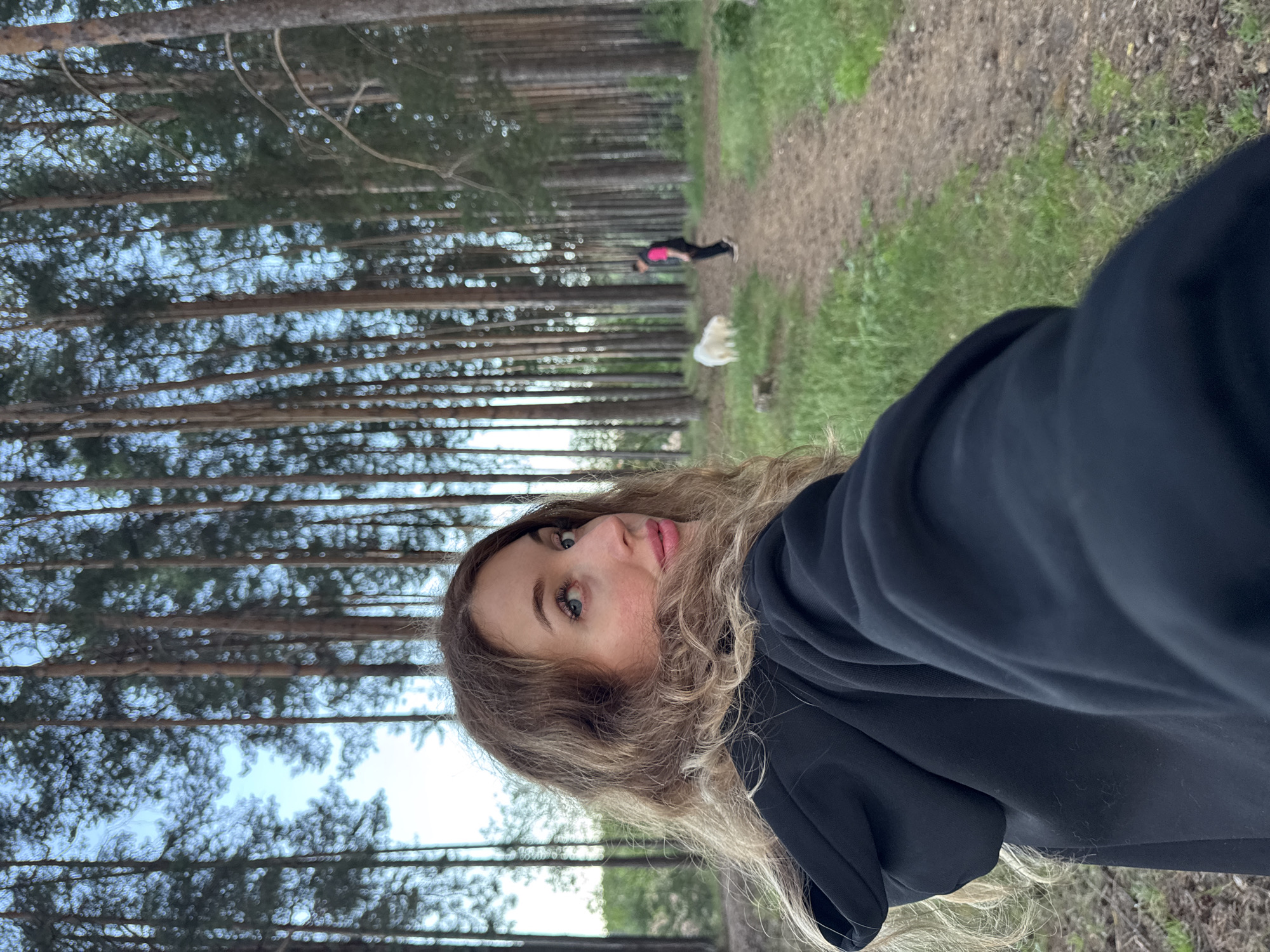A woman with long blonde hair is taking a selfie in a forest with tall pine trees. She is wearing a black hoodie. In the background, a person in a pink and black outfit is walking a white dog on a dirt path. The ground is covered with grass and pine needles. The sky is visible through the trees and appears to be cloudy. The location is the city of Chaikovsky, Russia.