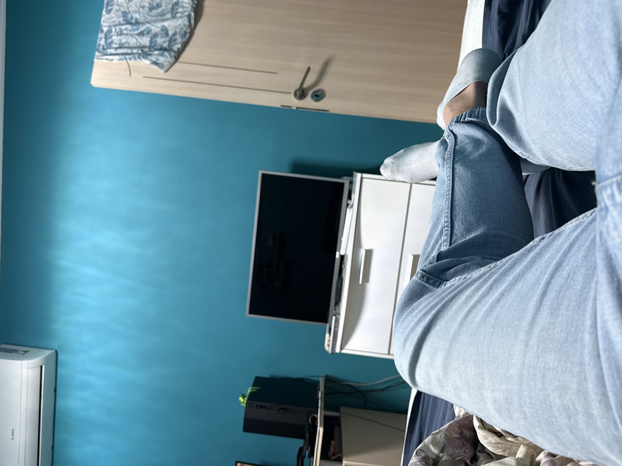 The photograph captures an interior setting with a notable focus on the seated individual. The person is wearing light-wash denim jeans and gray socks. A wardrobe with light-colored doors and metal handles is visible. Below the wardrobe, there is a black television screen and white storage drawers. Various electronic devices and cables are visible. The walls are painted in a teal color. Part of an air conditioning unit is visible at the bottom left corner. The location is in Varna, Bulgaria.