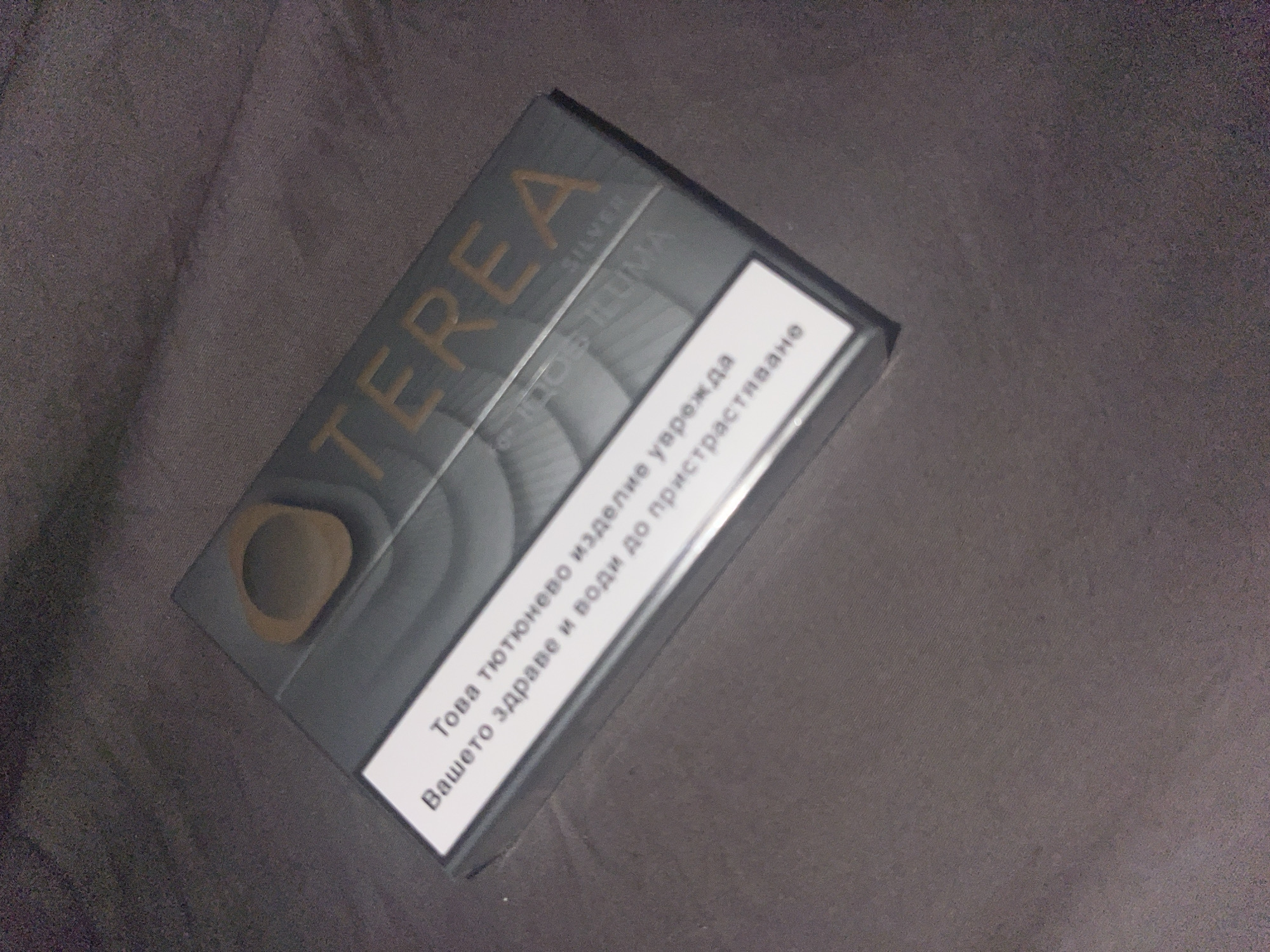 The image shows a close-up, angled shot of a silver and grey pack of Terea Silver IQOS ILUMA tobacco sticks. The pack is lying on a dark, textured surface that appears to be fabric, possibly a blanket or a couch. A white rectangular label with text in Bulgarian is prominently displayed on the front of the pack. The text translates to "This tobacco product harms your health and is addictive." The Terea logo is visible in orange-gold lettering on the upper part of the pack. The lighting is dim, casting shadows and creating a slightly grainy texture throughout the image. There are no people or animals present in the scene. The location is not specified in the image, and therefore cannot be determined to be Sofia, Bulgaria.