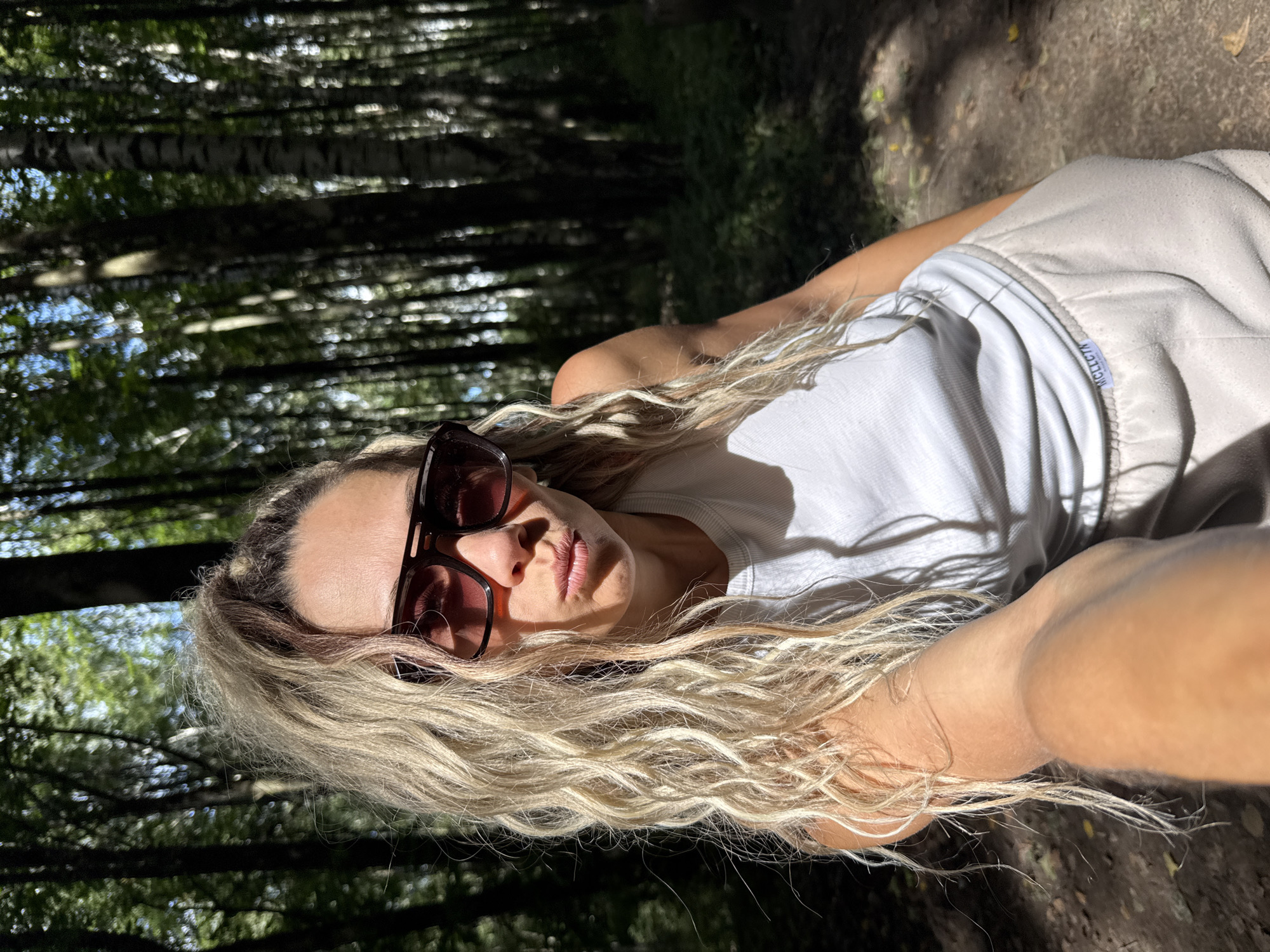 A woman with blonde curly hair, wearing sunglasses, a white tank top, and light-colored sweatpants, poses for a selfie in a forest. She is looking at the camera with her lips pursed. Sunlight streams through the trees, casting shadows on the ground. The location is described as Unknown, Unknown.