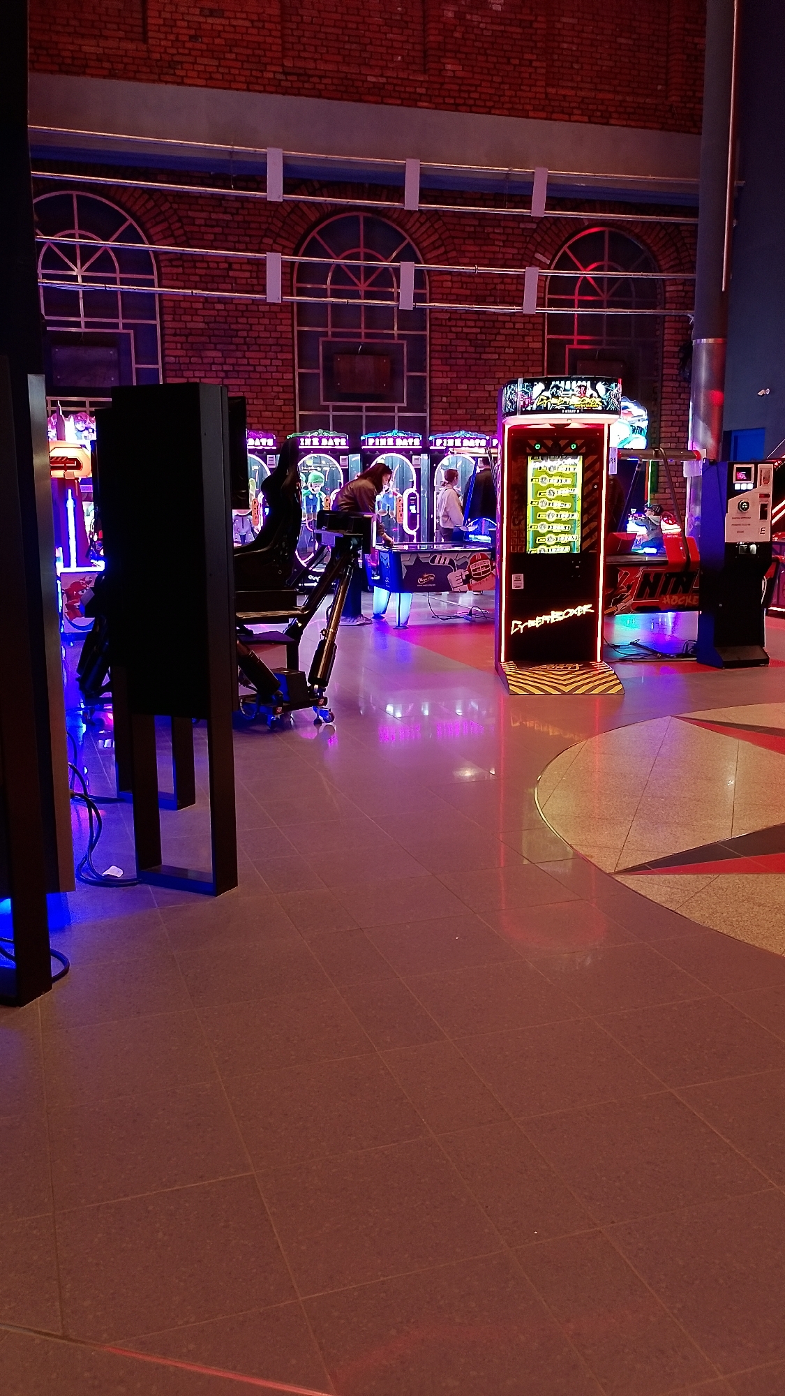 The image shows the interior of an arcade in Manufaktura, Łódź, Poland. Several arcade game machines are visible, including one labeled "Cyber Boxer". A few people are playing the games. The flooring consists of large tiles with a geometric star pattern. Arched windows and brickwork are prominent in the background, suggesting an industrial architectural style.