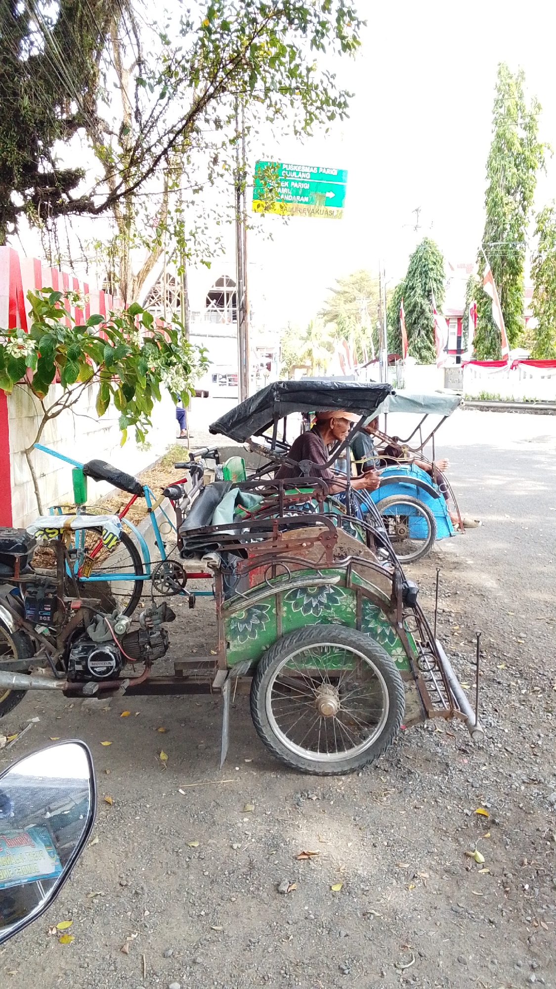 The image shows a roadside scene in Desa Karangbenda, Indonesia, with three pedicabs parked. The pedicabs have a motorcycle engine at the front and passenger seating at the back. Two men are seated in the pedicabs, one wearing a straw hat and the other in a dark shirt. The environment includes a gravelly roadside, trees, a building in the background, and a traffic sign. The sign has Indonesian text and indicates directions to PUSKESMAS PARIGI CUJILANG, TEK PARIGI, JAMU, and BANDARAN. The overall atmosphere suggests a typical day in a rural Indonesian town with pedicabs as a mode of local transportation.