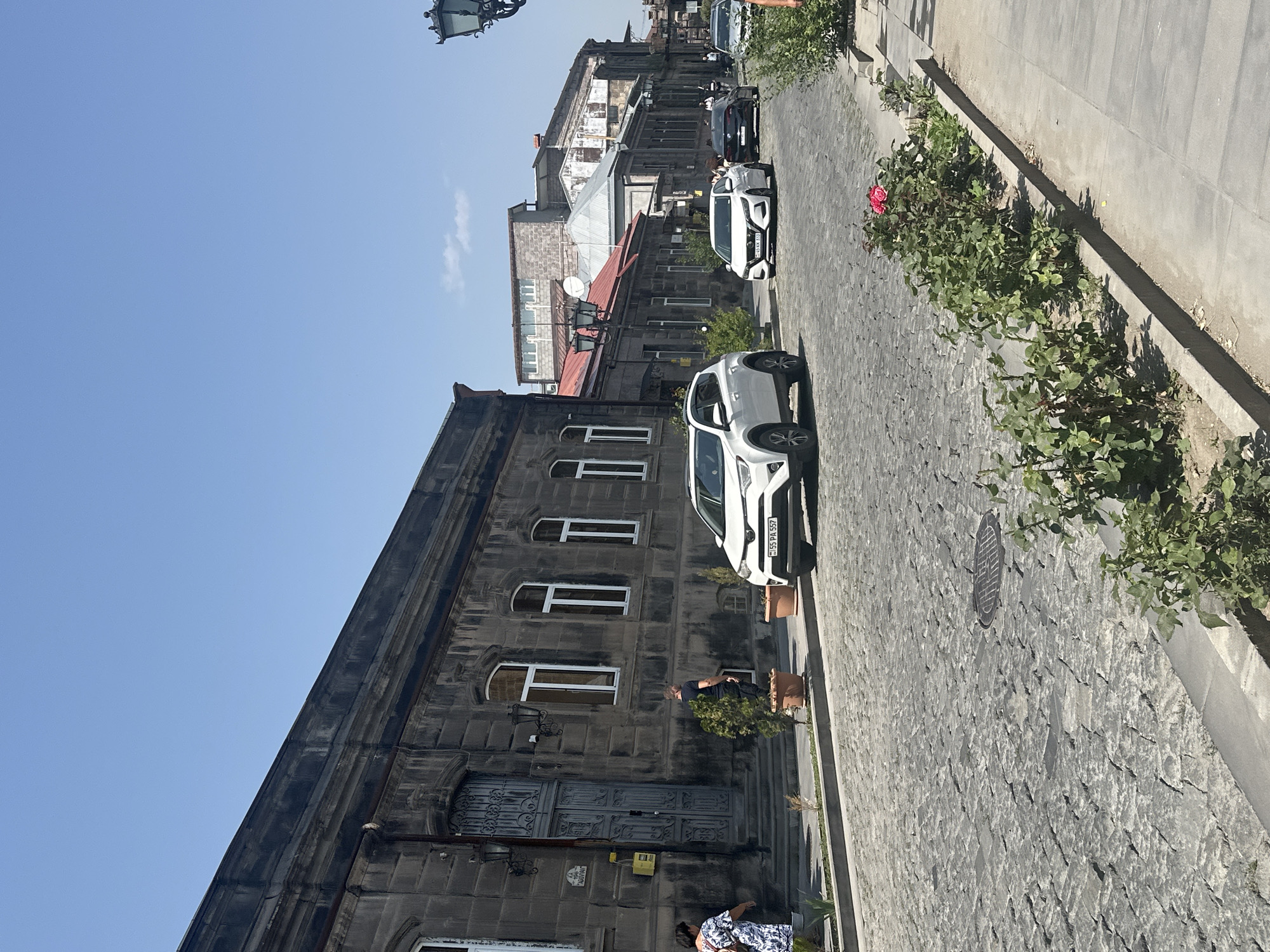 This is a street-level, eye-level, full-shot photograph taken in Gyumri, Armenia. The image captures a cobblestone street lined with stone buildings and a sidewalk with green bushes and flowering plants. Several cars are parked and driving on the street, including a white SUV, a white sedan, and a black sedan. Two people are visible on the sidewalk near the entrance of one of the buildings. The sky is clear blue, indicating a sunny day. The architecture of the buildings suggests a historical area within the city.