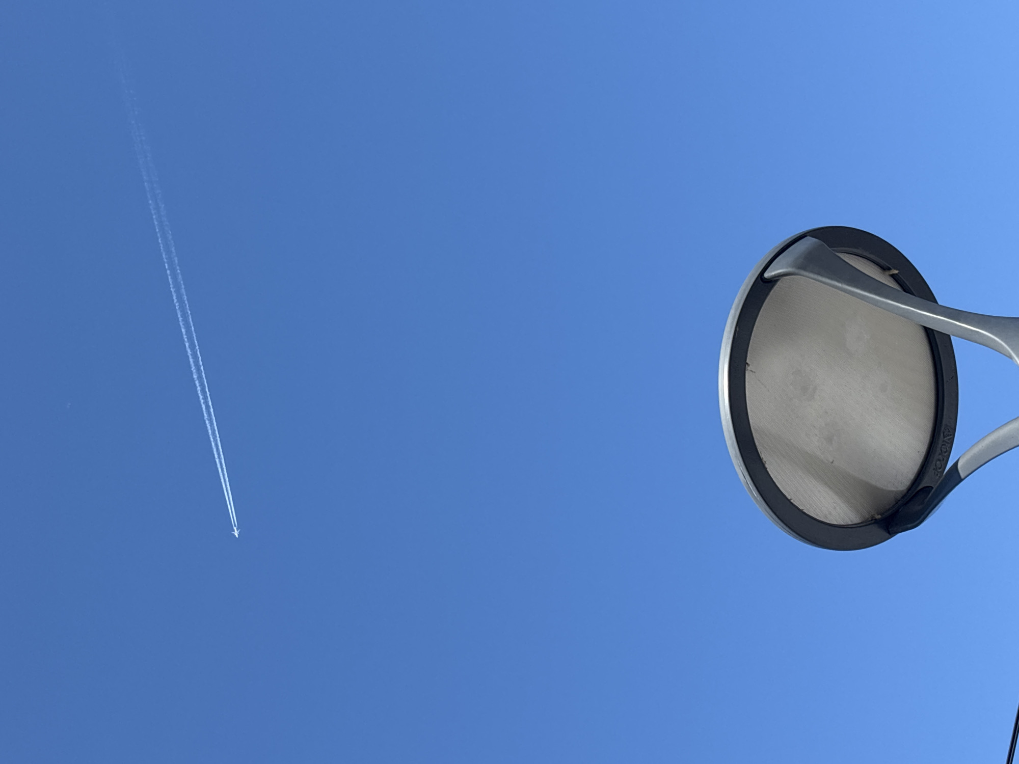 The image shows a bright blue sky with an airplane contrail stretching from the top-left to the center of the frame. A circular object, possibly a microphone pop filter, is located on the right side of the image. The object has a gray mesh center surrounded by a black ring, supported by a gray metal arm. The location is not provided.