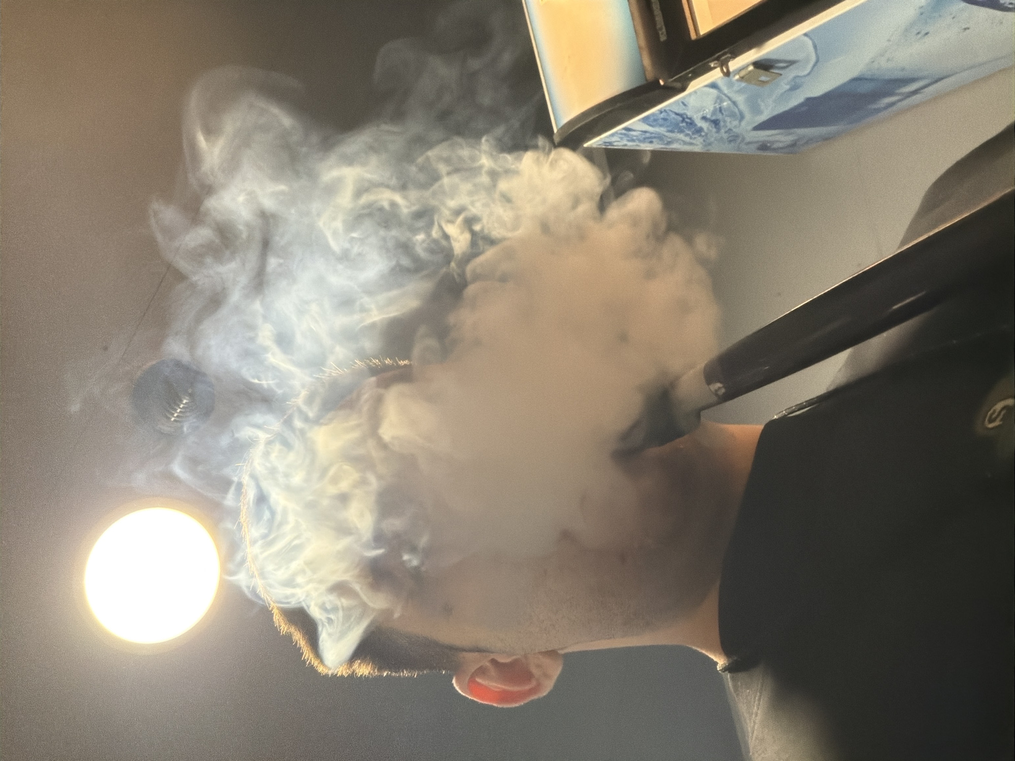 A man with short brown hair is exhaling a large cloud of white smoke. A dark tube extends from his mouth towards the top right of the frame, connected to an object with a light-colored top and a blue-and-white patterned side. A bright, circular light is visible to the left of the man's head. He is wearing a dark garment. The background is dark and out of focus. There is no evidence of the city of Varna, Bulgaria, in this image.