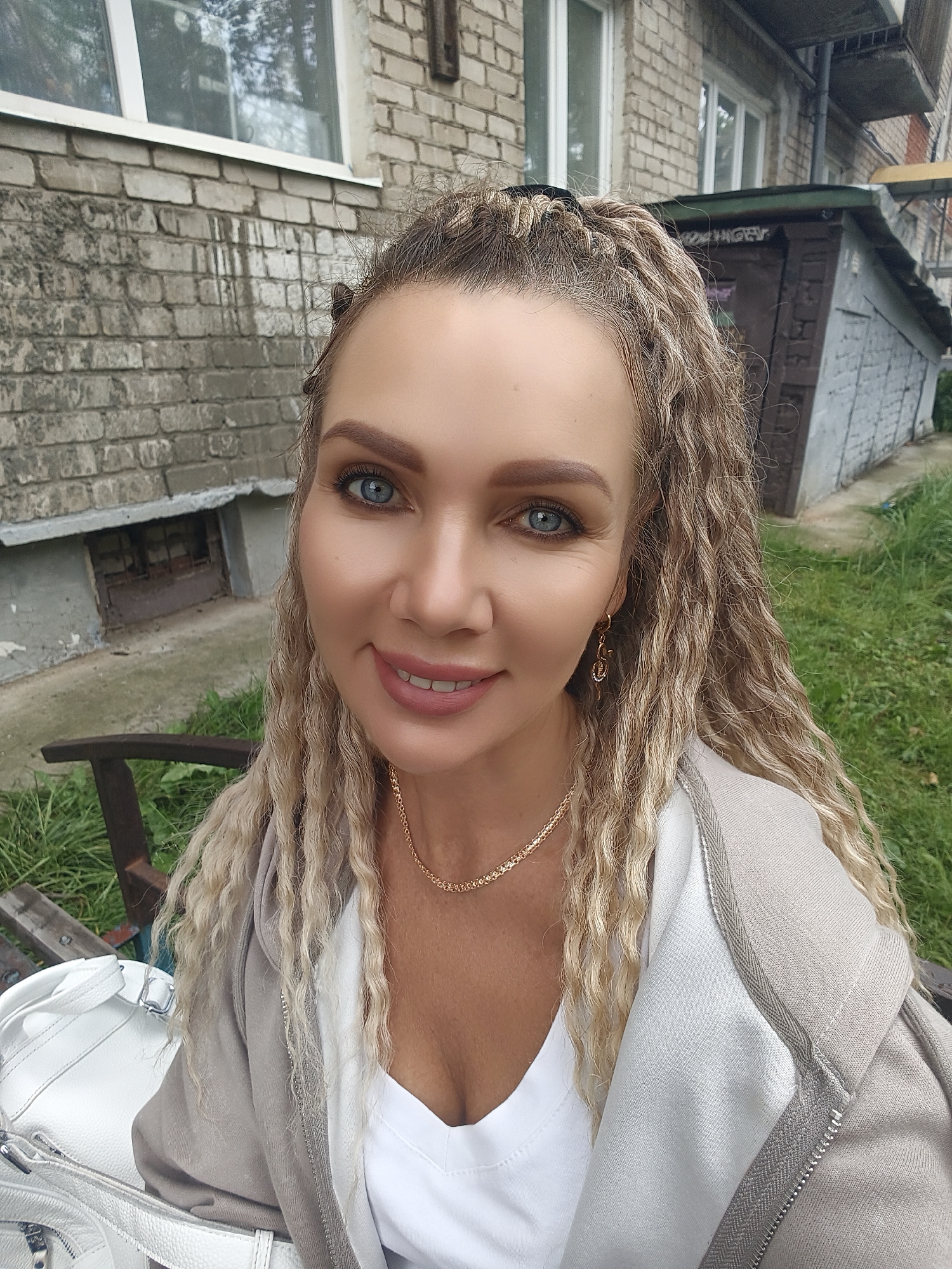 A woman is posing for a selfie in front of a brick building and some greenery. The woman has long, curly blonde hair and blue eyes. She is wearing a white shirt and a light-colored hooded jacket. A white bag is visible in the foreground, and a wooden bench is behind her. The building appears to be an apartment building, with windows visible on the upper floors and a small, barred window on the ground floor. A grey, garage-like structure is to the right of the building. The overall scene suggests an outdoor setting in a somewhat urban or suburban environment.
