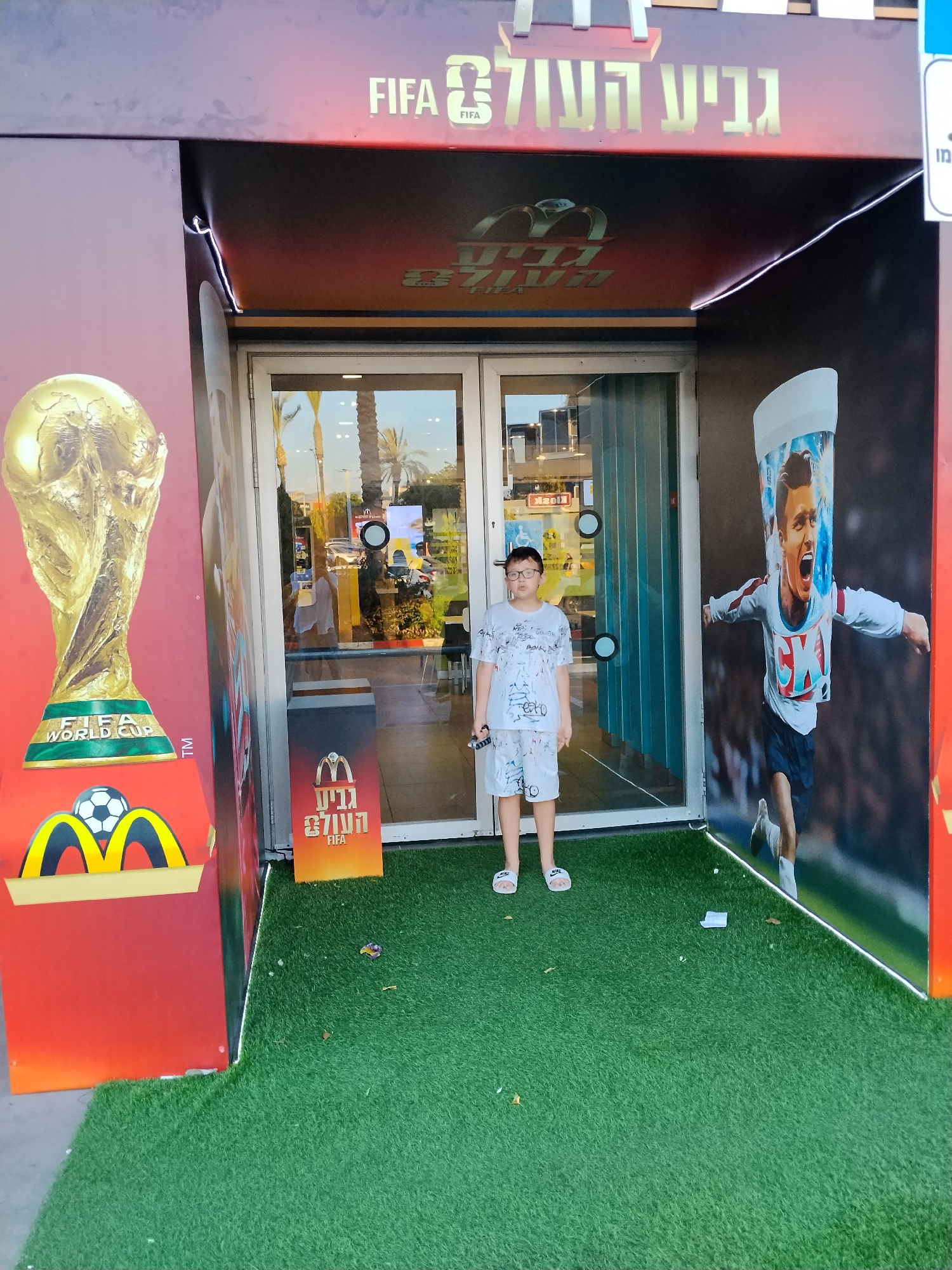 A young boy stands on a patch of artificial grass in front of a McDonald's restaurant entrance. The McDonald's appears to be decorated with FIFA World Cup themes, featuring a large image of the FIFA World Cup trophy and a mural of a soccer player. The boy is wearing a white t-shirt and shorts with a graffiti-like pattern and white slides. He is holding a small black object in his right hand. The entrance to the restaurant has glass doors and windows, and palm trees are visible in the background through the glass. The text "FIFA" and Hebrew text, which translates to "The World Cup," are prominently displayed on the facade of the building. The image is taken outdoors during daylight. The location is identified as Be'er Sheva, Israel.