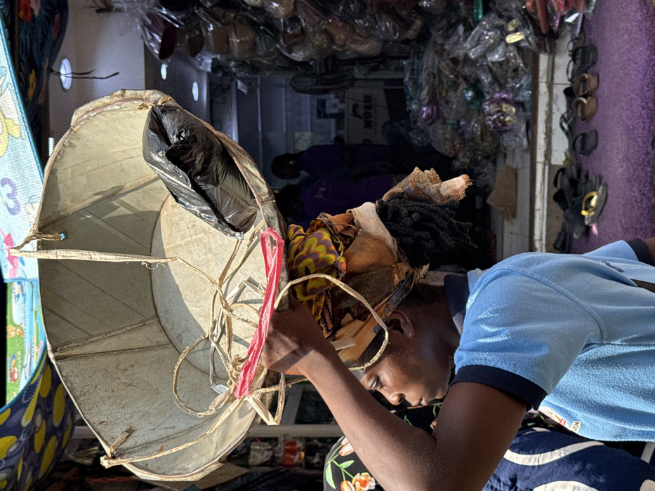 The image depicts a bustling outdoor market scene in Lagos, Nigeria. A young woman, viewed in profile facing left, carries a large, weathered, silver-colored metal basin on her head. Her right hand is raised, holding the edge of the basin, which contains a black plastic bag and is secured by ropes and a red strap over a patterned fabric head-wrap. She wears a light blue polo shirt with dark blue trim on the collar and sleeves. In the mid-ground, a retail shop with a darker interior is visible, brightly lit by internal lights. Two indistinct figures in dark, possibly purple, clothing are present within the shop. The shop's display area is densely packed with numerous items, appearing to be shoes or bags, individually packaged in clear plastic. On the ground directly outside the shop, a purple mat is covered with various sandals and flip-flops. A partially obscured sign inside the shop reads "NORM." An overhead colorful awning, patterned with blue and yellow, is visible at the top, indicating a sunny outdoor market environment.