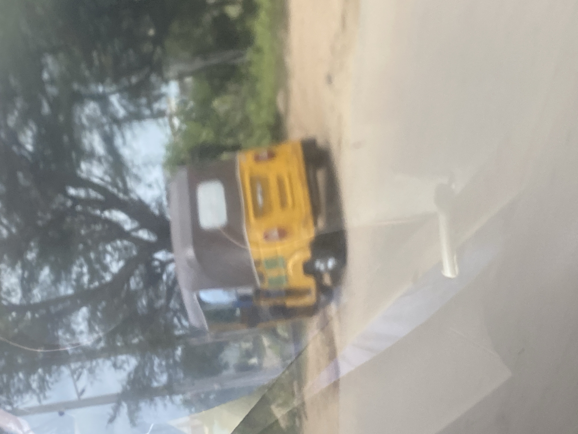 The image depicts an auto rickshaw, likely a Bajaj RE or similar model, driving on a road in Karmajiji, Nigeria. The auto rickshaw is predominantly yellow with a brown or grey roof. The background features trees and vegetation. The photo is taken through a window, possibly from inside another vehicle, as evidenced by the blurry reflection and white interior panel.