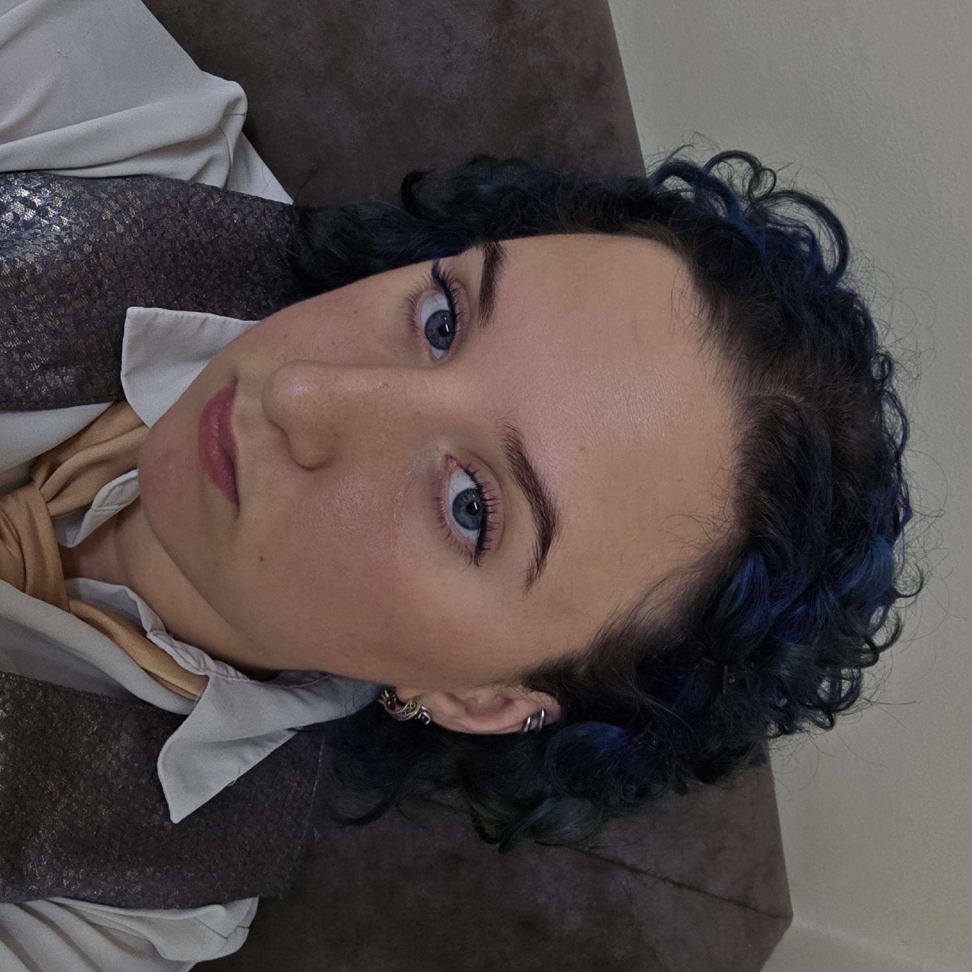 This is a close-up, head-and-shoulders portrait of a young person with curly, dark hair with blue highlights. The individual has blue eyes, a fair complexion, and is wearing makeup including mascara and eyeliner. They are dressed in a white collared shirt, a gold scarf tied around the neck, and a dark, textured vest. The person is wearing earrings on their left ear, including a hoop and a decorative earring. The background appears to be an indoor setting with a dark, textured surface, possibly upholstery. There is no discernible indication of the city of Chișinău, Moldova.