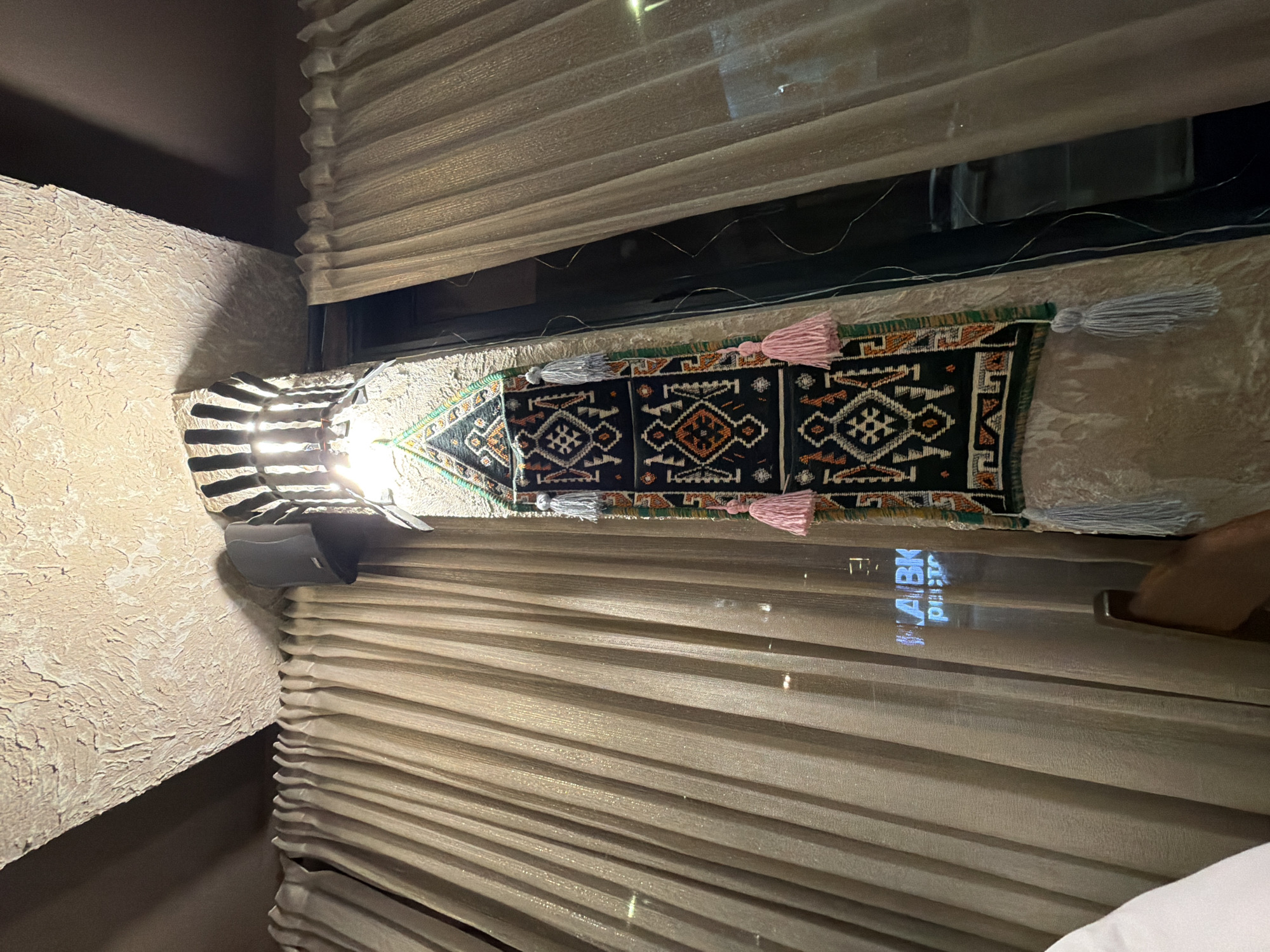 The image shows a decorative tapestry hanging from a pillar in what appears to be an indoor establishment. The tapestry is dark with intricate patterns in white, orange, and green, and features several tassels, two of which are pink and two are light gray. Above the tapestry, a light fixture with vertical slats illuminates the area. A dark gray speaker is mounted on the pillar above the light. The pillar itself is textured, resembling rough stone. To the left and right of the pillar are large, pleated curtains, light beige in color, with a subtle shimmer. Through a window or opening behind the tapestry and curtains, a reflection of blue neon lights spelling out "HABK" and "РИСТО" is visible, suggesting the presence of a sign from across the street or within the establishment. A portion of a white garment and a wooden chair are visible in the lower left and lower right of the frame, respectively. The ceiling above the pillar is also textured and light beige. The scene is indoors, with artificial lighting. The location is identified as Kumbor, Montenegro.