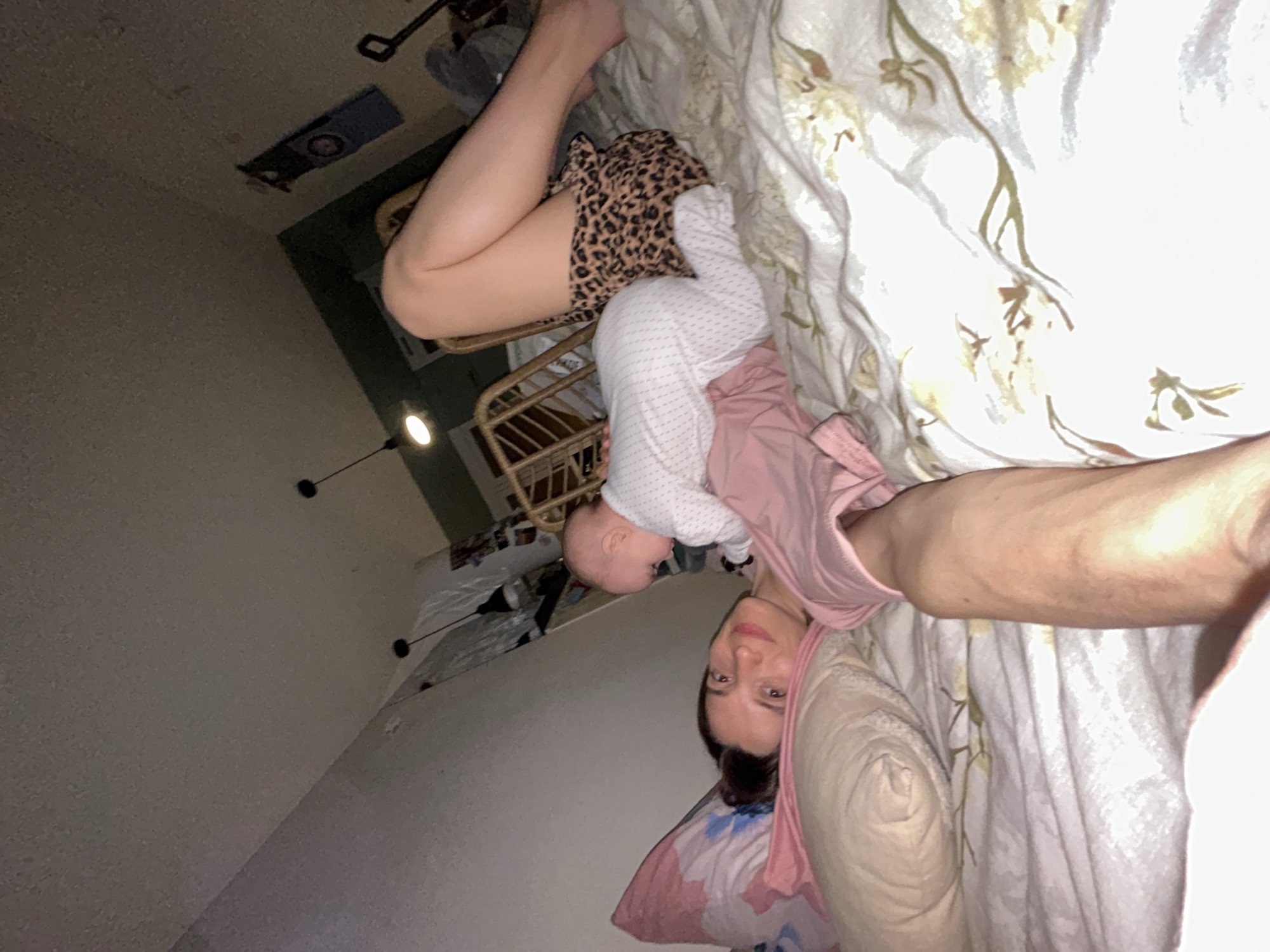 A woman lies in bed with a baby, taking a selfie. The woman wears a pink shirt and rests her head on a pink and white patterned pillow against a white floral-patterned bedsheet. The baby, dressed in white with a pink top, is positioned near her legs which are clad in a leopard-print short. The bed is placed next to a baby cot. In the background, a room wall stretches to the left. A lighting fixture hangs on the left wall. It is impossible to determine if the location is Moscow, Russia from this image alone.