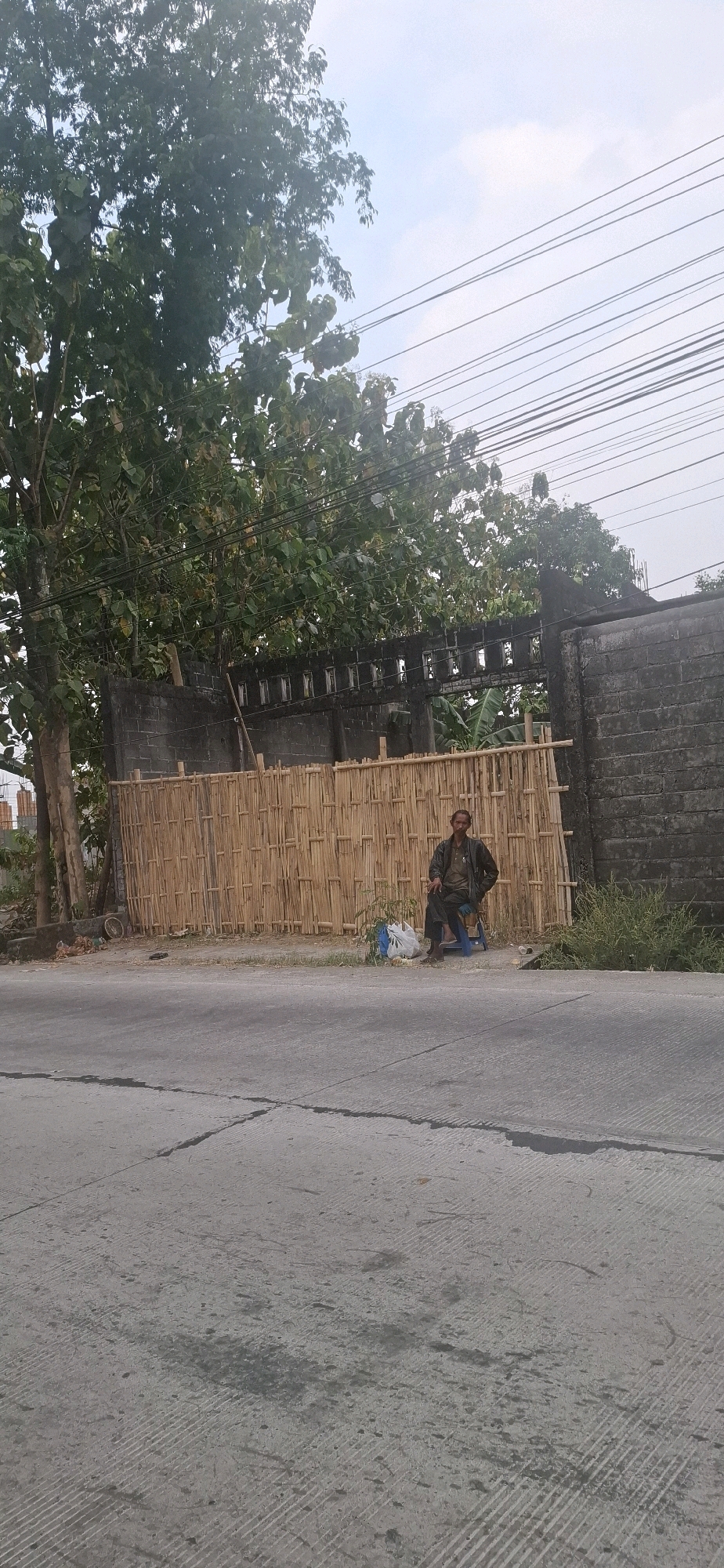 A man sits on a blue plastic chair next to a tall bamboo fence in Desa Gagaksipat, Indonesia. The man is wearing a dark jacket and pants. He is holding a cigarette in his right hand and looking to his left. A white plastic bag and a small plant are on the ground next to him. Behind the bamboo fence is a grey concrete wall and trees. Power lines are visible in the sky above. The scene is on the side of a paved road.
