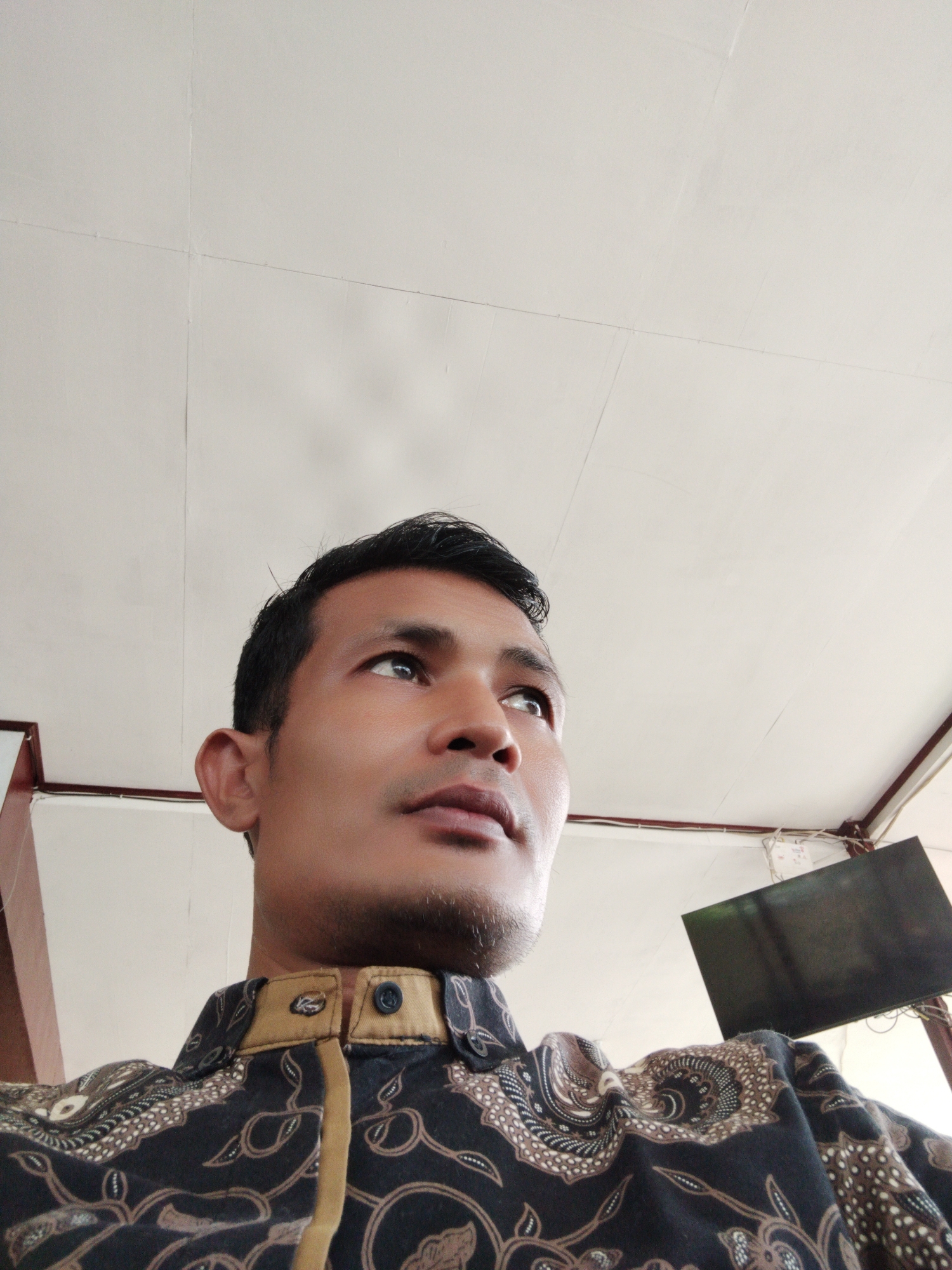 The image presents a low-angle, close-up portrait of a man looking upwards and towards the right side of the frame. The subject is an adult male, likely of Southeast Asian descent, with short, dark hair and light stubble on his chin and jawline. He is wearing a dark-colored, long-sleeved shirt with an intricate, ornate pattern in brown and cream tones, characteristic of batik fabric. The shirt features a standing collar with tan or gold-colored trim and visible buttons.
The man's expression is neutral and focused, suggesting a moment of observation or contemplation of something above or in the distance. His eyes are open and directed away from the camera.
The background primarily consists of a light-colored, possibly white or off-white, ceiling with visible seams, indicating ceiling panels or a painted surface. Dark-colored structural beams or architectural elements are visible along the left and right edges of the frame. In the upper right corner, a dark, rectangular object, resembling a mounted television or monitor, can be seen, accompanied by some visible wires or cables.
The setting is indoors, with ambient lighting that does not strongly indicate a specific time of day or outdoor weather conditions. The scene is located in Lhokseumawe, Indonesia. No discernible text is visible within the image.