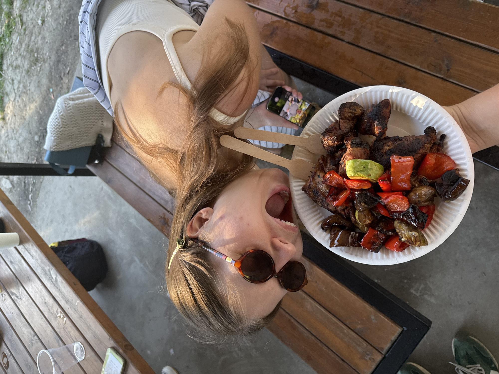 A young woman with long brown hair is holding a paper plate full of grilled meat and vegetables in Cukarica Urban Municipality, Serbia. She is lying on a wooden bench, wearing sunglasses and a white tank top with a striped shirt over it. Her mouth is open as if she is about to eat the food. Next to her on the bench is a smartphone and a white sweater. Under the bench is a black bag and a wooden table. The plate contains what appears to be grilled meat, red peppers, zucchini, and eggplant. The photo is taken at a tilted angle.