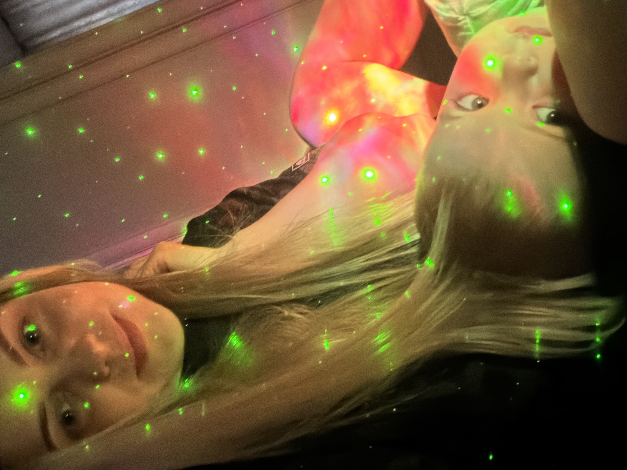 This image captures two individuals, likely women with light blonde hair, in an indoor setting illuminated by a vibrant light projector. The scene is dominated by numerous small, bright green laser-like dots scattered across the frame, resembling stars or fireflies, and larger, amorphous red and orange light patterns projected onto the subjects and background, creating a dreamy or party-like atmosphere.
The image is rotated, presenting the subjects as if lying on their sides. The person in the foreground is closer to the viewer, with light blonde hair fanned out. Their face is clearly visible, featuring a gentle, slightly amused smile as they look towards the camera. The green dots highlight features on their face and hair.
Further back and slightly above, another person is partially visible. They also have light blonde hair and are looking upwards with a neutral, possibly curious or surprised expression. Their body and face are similarly bathed in the green and red/orange projected lights.
The background appears to be a wall or ceiling, also covered in the light projections. The overall impression is one of a relaxed, intimate moment captured in a playfully lit environment, likely in the evening or at night. No specific location cues beyond an indoor setting are visible, nor is any text. The location context provided is Votkinsk, Russia.