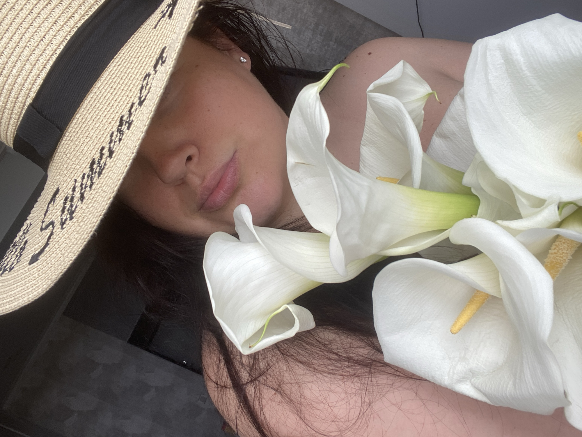A woman wearing a straw hat with a black ribbon and script text on the brim is holding a bouquet of white calla lilies. The hat partially obscures her face, revealing her nose and lips. Her dark hair falls around her shoulders. She is wearing a small silver stud earring in her visible ear. The background is dark and appears to be indoors, possibly a room with a textured wall and a window or doorway. The image is a close-up shot, focusing on the woman and the flowers. The location is not specified as Sochi, Russia.
