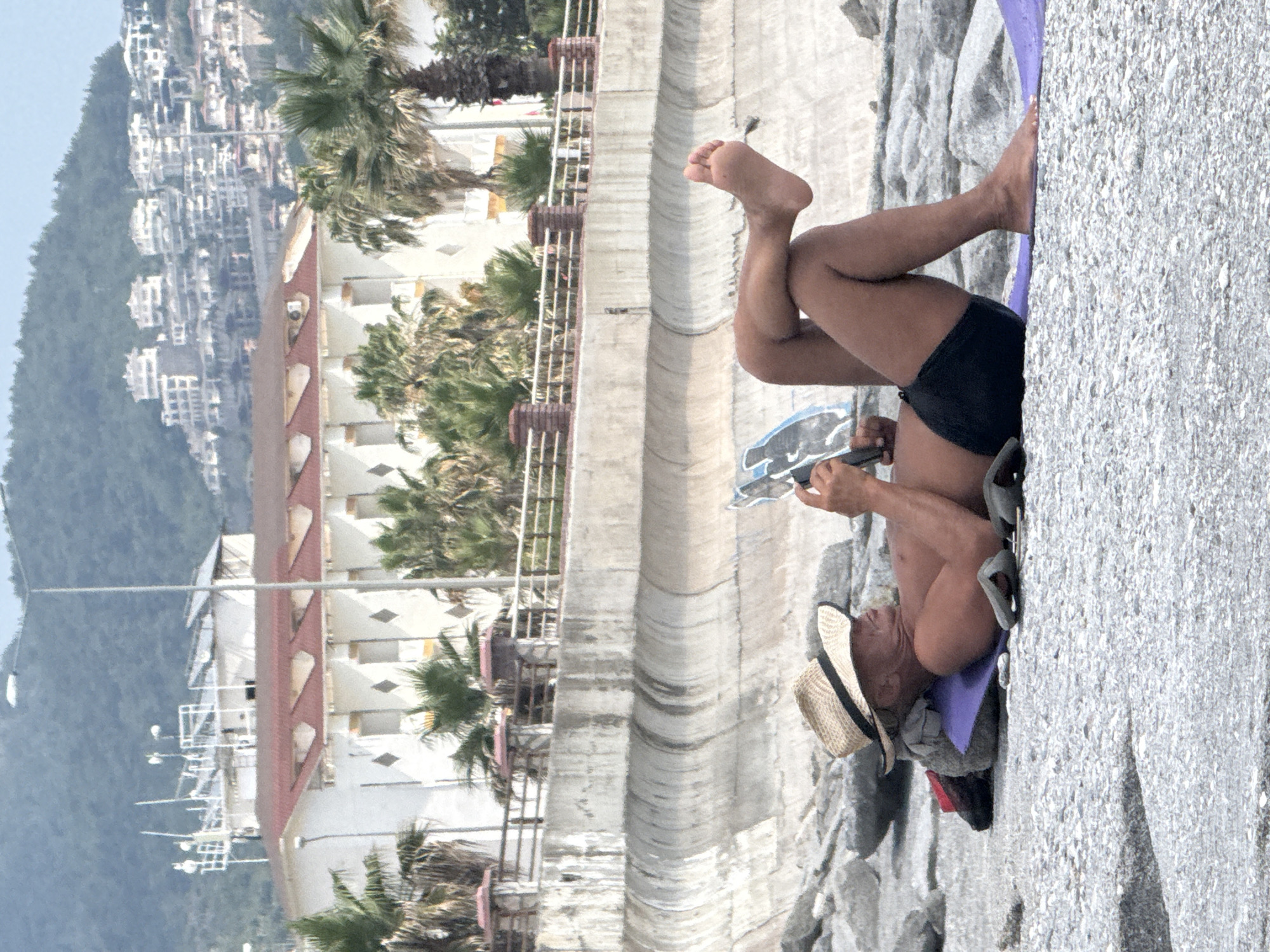 A man wearing a straw hat and dark swim trunks lies on his back on a purple mat on a rocky beach, reading a magazine. His legs are crossed, and his right foot is raised. Behind him is a concrete seawall, and further back, buildings and palm trees are visible on a hillside. The scene appears to be in Alanya, Turkey, given the architecture and vegetation often found there.