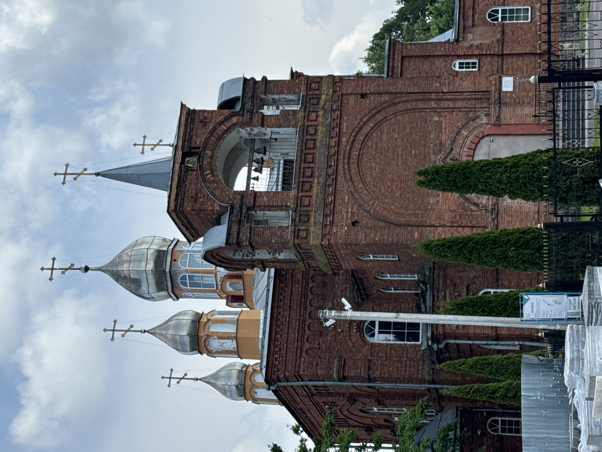 The image shows the Church of St. Nicholas in Chudovo, Russia. The church is made of red brick and features several domes topped with crosses. The central structure includes a bell tower with an arched opening containing bells. The church has numerous arched windows, and the grounds are surrounded by a black metal fence. In the foreground, there are stacks of building materials, including what appear to be paving stones wrapped in plastic. Several tall, conical evergreen trees are planted along the side of the church. The sky is cloudy.