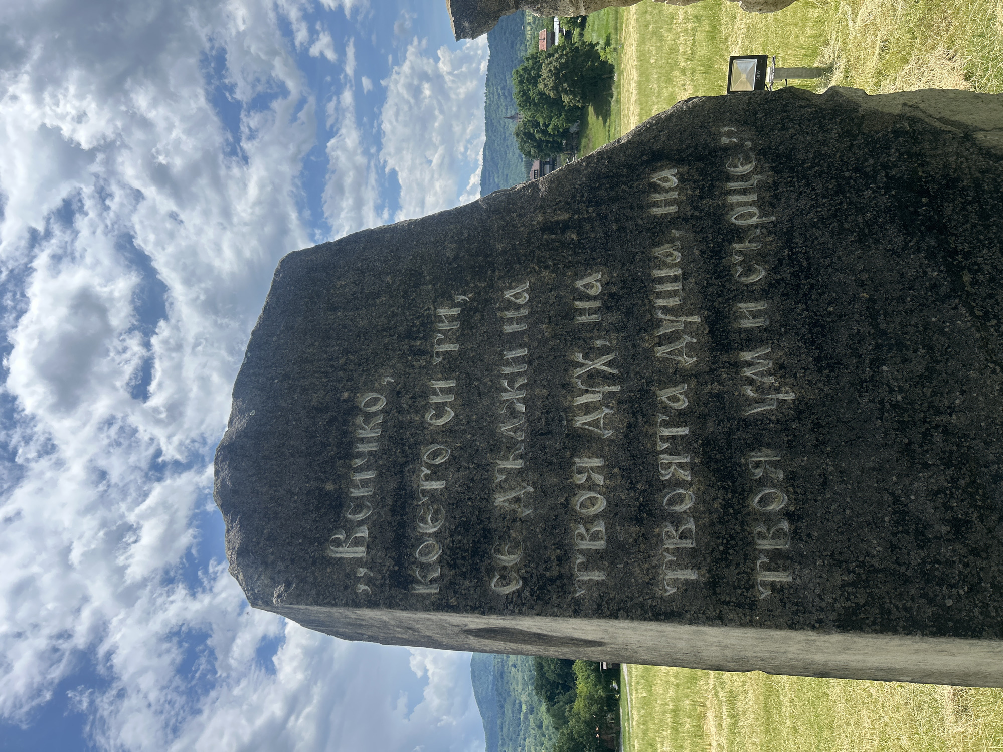 A large, dark grey stone monument, engraved with text in Cyrillic, stands prominently in the foreground. The text on the stone, translated from Bulgarian, reads: "Everything you are is due to your spirit, to your soul, to your mind and heart." The letters are carved into the rough surface, appearing lighter against the darker stone.
The setting is an outdoor area, likely a park or memorial site, with a vast, partly cloudy sky visible above. White, wispy clouds are scattered across a bright blue sky, indicating fair weather during daytime. In the mid-ground and background, distant green mountains rise, covered with lush vegetation and trees, suggesting a mountainous or hilly region. A few scattered buildings are visible among the trees at the foot of the mountains, hinting at a small settlement in the valley.
The ground surrounding the monument is covered in vibrant green grass. To the side of the stone, a black floodlight, labeled "30W," is visible, mounted on a stand. The scene evokes a sense of tranquility and contemplation, characteristic of a commemorative site in a natural, scenic landscape, consistent with a location like Gardevtsi, Bulgaria.