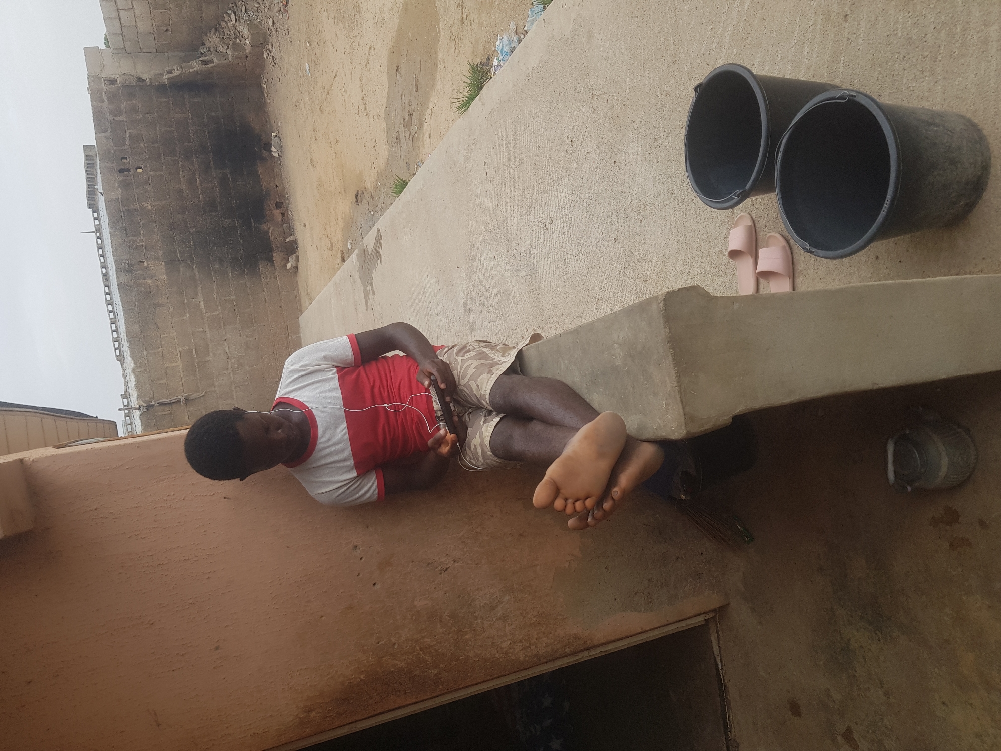A man is reclining on a concrete structure outdoors in what appears to be Dutsinma B, Nigeria. He is wearing a red and white shirt, patterned shorts, and has earphones plugged in. He is looking at a phone he is holding. Near him are two black buckets, a pair of pink sandals, and a small metal container. A brick building is in the background, and the sky is overcast.