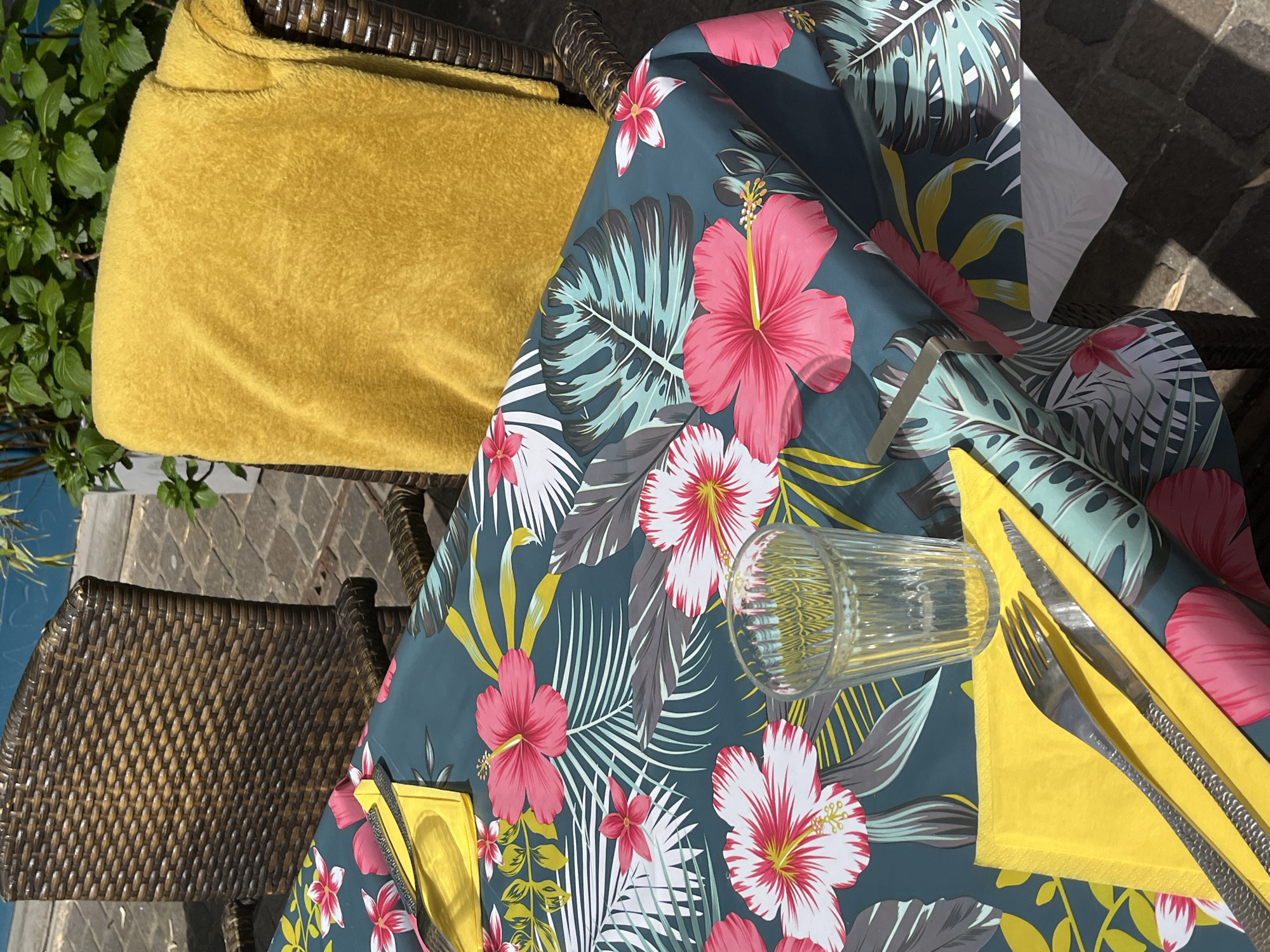 A high-angle shot captures a table setting outdoors. The table is covered with a dark teal tablecloth adorned with a tropical floral pattern featuring pink hibiscus and various stylized leaves. A yellow-gold folded napkin is present, topped with a fork and knife. Next to the napkin is a clear, empty drinking glass lying on its side.
A portion of a wicker chair is visible, draped with a yellow-gold towel or blanket. The background includes a view of stone paving and greenery.
The image appears to be taken in Metz, France, based on contextual clues.