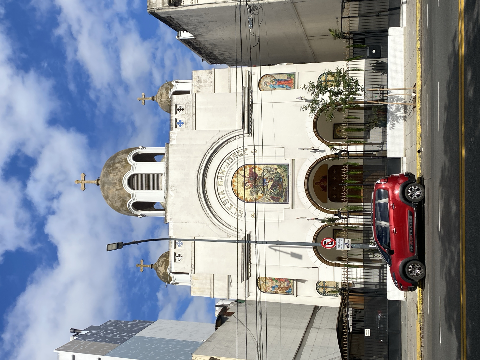 Here is a detailed description of the image:
The image captures a white Orthodox church located in the city of Buenos Aires, Argentina. The church showcases a distinctive architectural style with domes topped with crosses. A mosaic depicting a religious scene is positioned above the entrance arches. The name “IGLESIA SAN JORGE” (Saint George Church) can be read on the building's facade.
A red Citroen Aircross is parked at the side of the street, partially obscuring the view of the church's entrance. A streetlamp and power lines cut across the front of the church. A traffic sign denoting parking regulations is also visible. The sky behind the church is bright blue with scattered clouds. The buildings beside the church appear to be modern structures, which creates a contrast with the church’s traditional design.