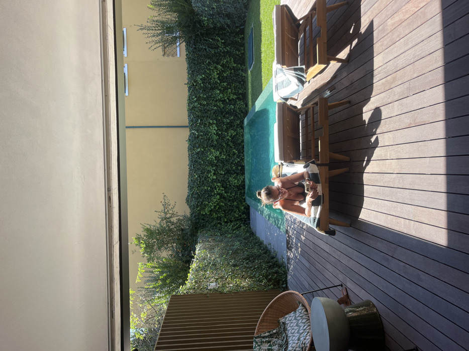 An overhead view captures an outdoor residential patio in Lordelo do Ouro e Massarelos, Portugal, featuring a swimming pool and a sunbathing area. The foreground displays dark composite wood decking. On the left, a wicker armchair with a green and white patterned cushion and blanket sits next to a cylindrical concrete side table. In the mid-ground, a female individual, wearing a dark bikini and eyeglasses with her hair in a bun, lies prone on her stomach on a wooden sun lounger, holding and looking at a smartphone. A white and dark green striped towel is draped over her lounger. Two additional empty wooden sun loungers, one with a similar striped towel, are positioned to her right. Beyond the loungers, a rectangular swimming pool with turquoise water occupies the center-rear. The pool is bordered by green, dense ivy-covered walls or hedges. Behind the vegetation, a tall, plain yellow building wall is visible, featuring two small rectangular windows on its upper right section. Sunlight illuminates the pool and sun loungers, casting shadows on the decking. A vertical slatted privacy screen runs along the far left edge of the patio.