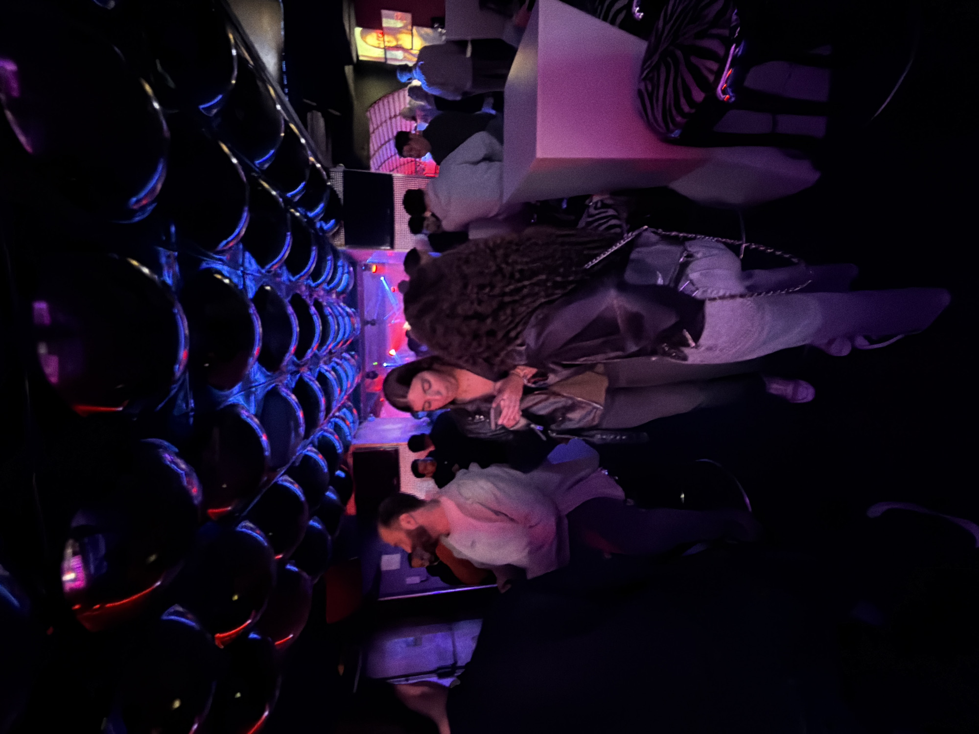The image shows an indoor scene at what appears to be a nightclub or bar, based on the low lighting, interior design, and the presence of a group of people. The image is slightly rotated.
People: A group of people are present. There is one woman with dark curly hair wearing a jacket and light-colored pants, possibly jeans. Another person, a man, appears to be wearing a light-colored shirt and darker pants and has a beard. Other people are visible in the background.
Objects: There are several notable objects. A wall with a textured design, resembling a pattern of dark spheres with blue and red lighting, is on the left. A table or seating area with a pink-lit surface is visible in the background.
Actions/Interactions: The individuals appear to be socializing, possibly engaged in conversations or relaxing in the club environment.
Environment: The setting is dark with artificial lighting that creates a nightclub or bar ambiance. The background includes additional architectural elements, furniture, and lighting.
Location: While the image mentions Vila Real, Portugal, there is no visual information that enables the identification of an exact location within the city. Therefore, the physical coordinates for Vila Real are: 41.3000° N, 7.7500° W.