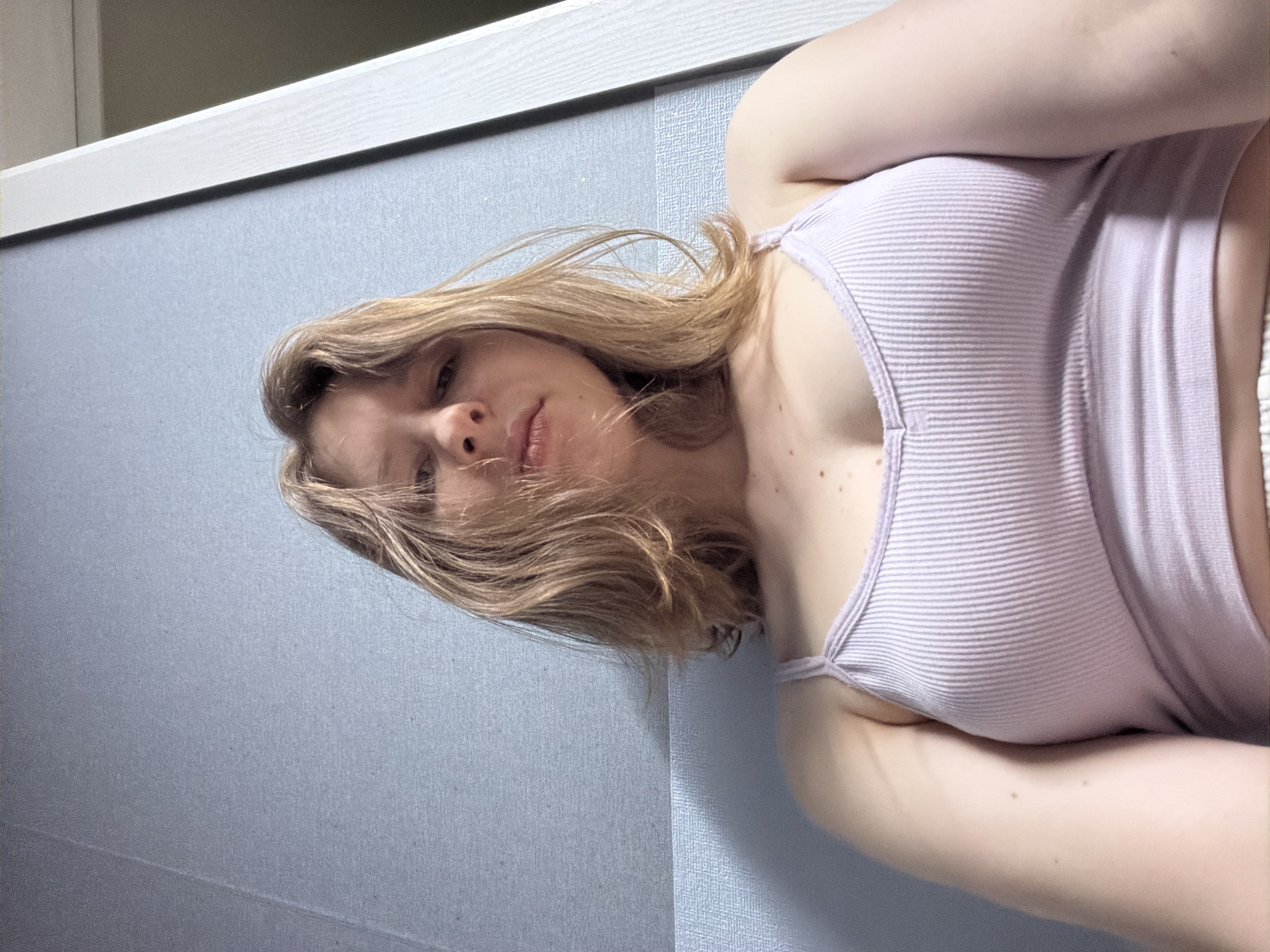 A young woman with blonde hair is lying down and looking up at the camera. She is wearing a light purple ribbed tank top. Her hair is spread around her head. The background is a blue wall with a white trim at the top. The lighting suggests it is daytime. The woman appears relaxed. There is no visible text in the image.