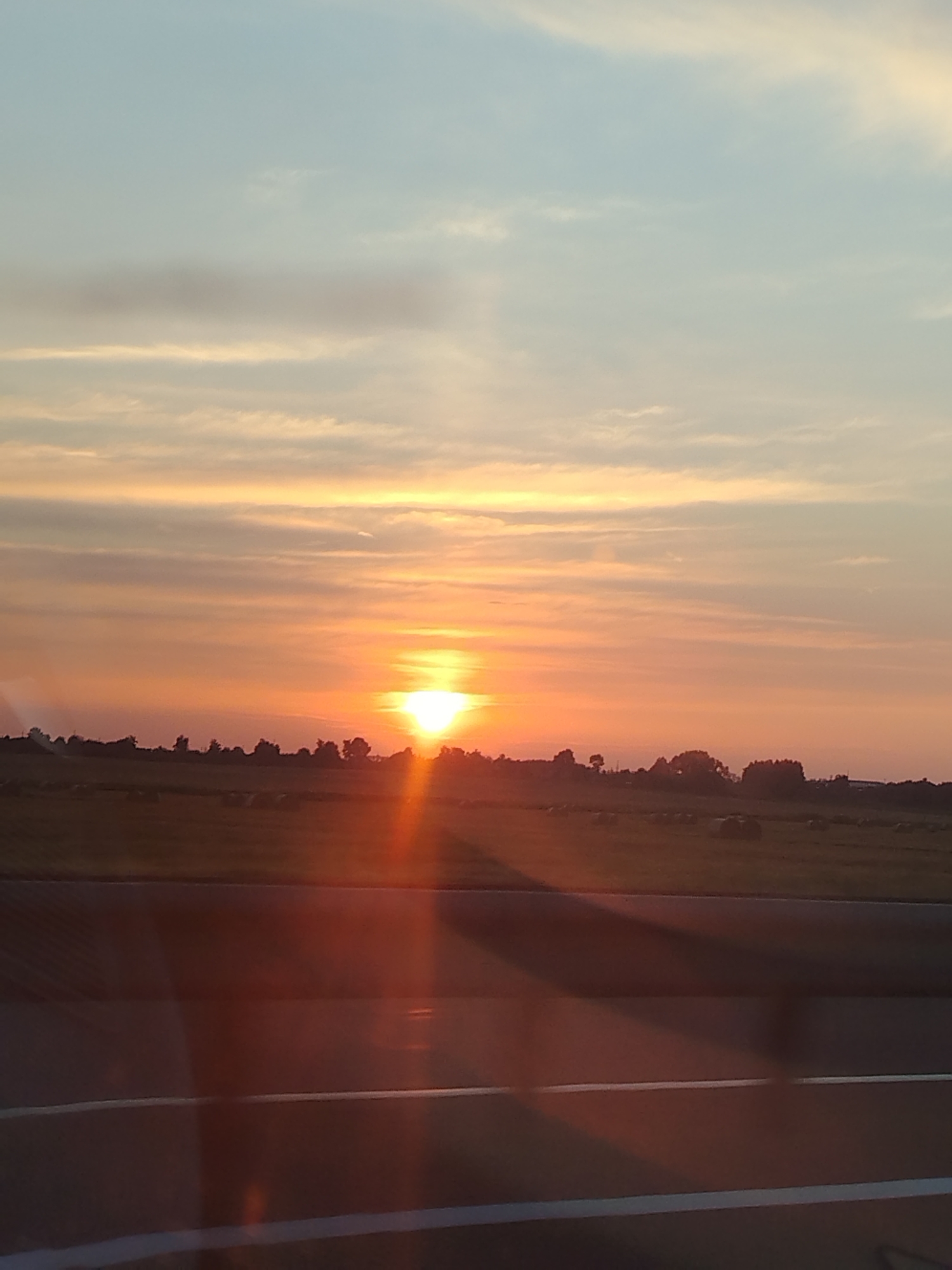 The image shows a sunset over a rural landscape. The sun is low on the horizon, casting a warm orange glow across the sky. There are a few clouds in the sky, which are tinged with pink and gold. In the foreground, there is a field with hay bales scattered across it. There are also some trees in the distance. The overall impression is one of peace and tranquility.