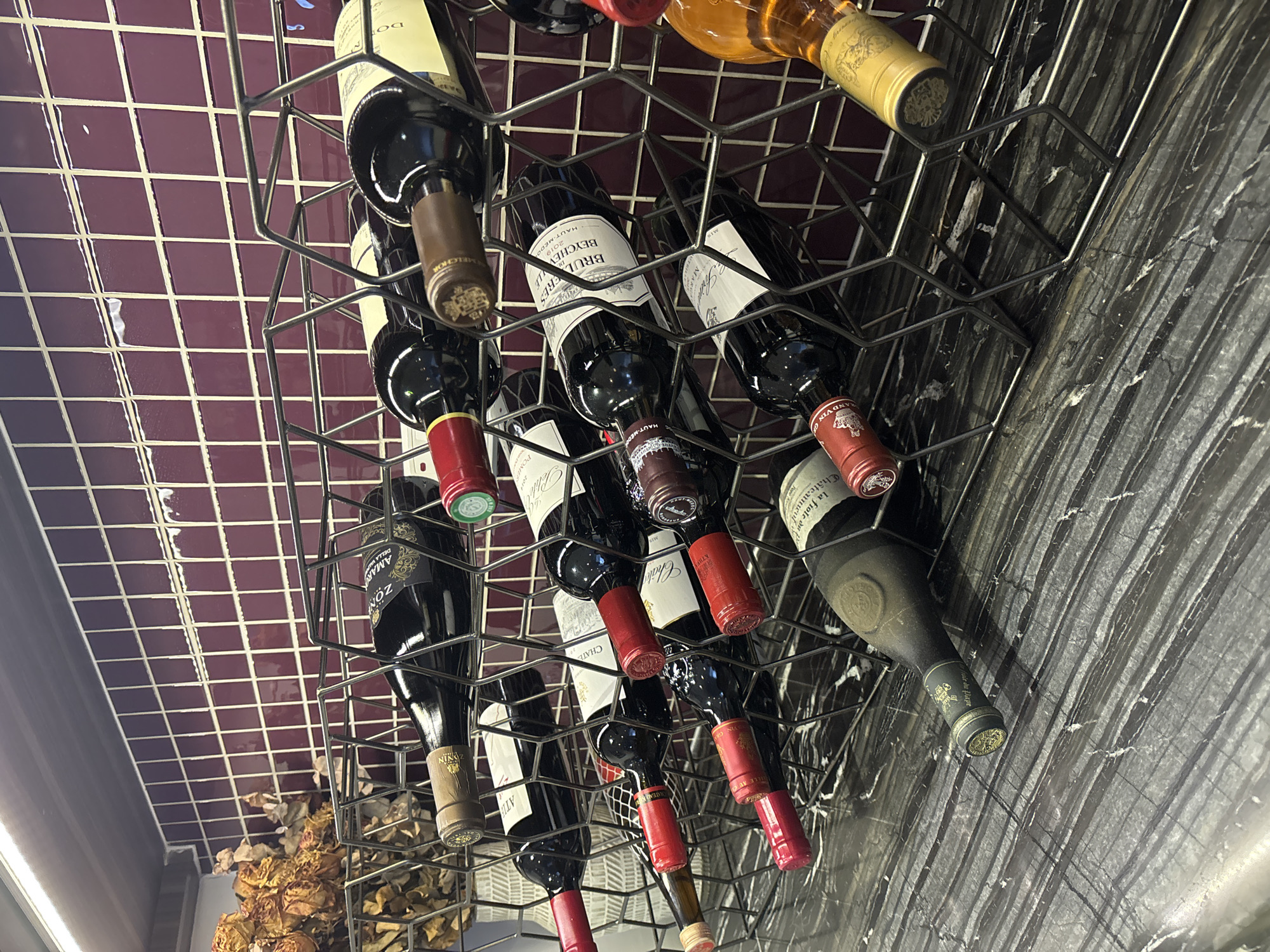 A wine rack holding multiple bottles of wine is placed on a black marble countertop in front of a burgundy tiled wall. The wine rack is a tiered metal structure with a geometric design. Some bottles are upright, while others are angled. The countertop has a prominent veined pattern. Dried flowers are visible in a basket to the left of the wine rack. The scene appears to be indoors, possibly in a restaurant or home.